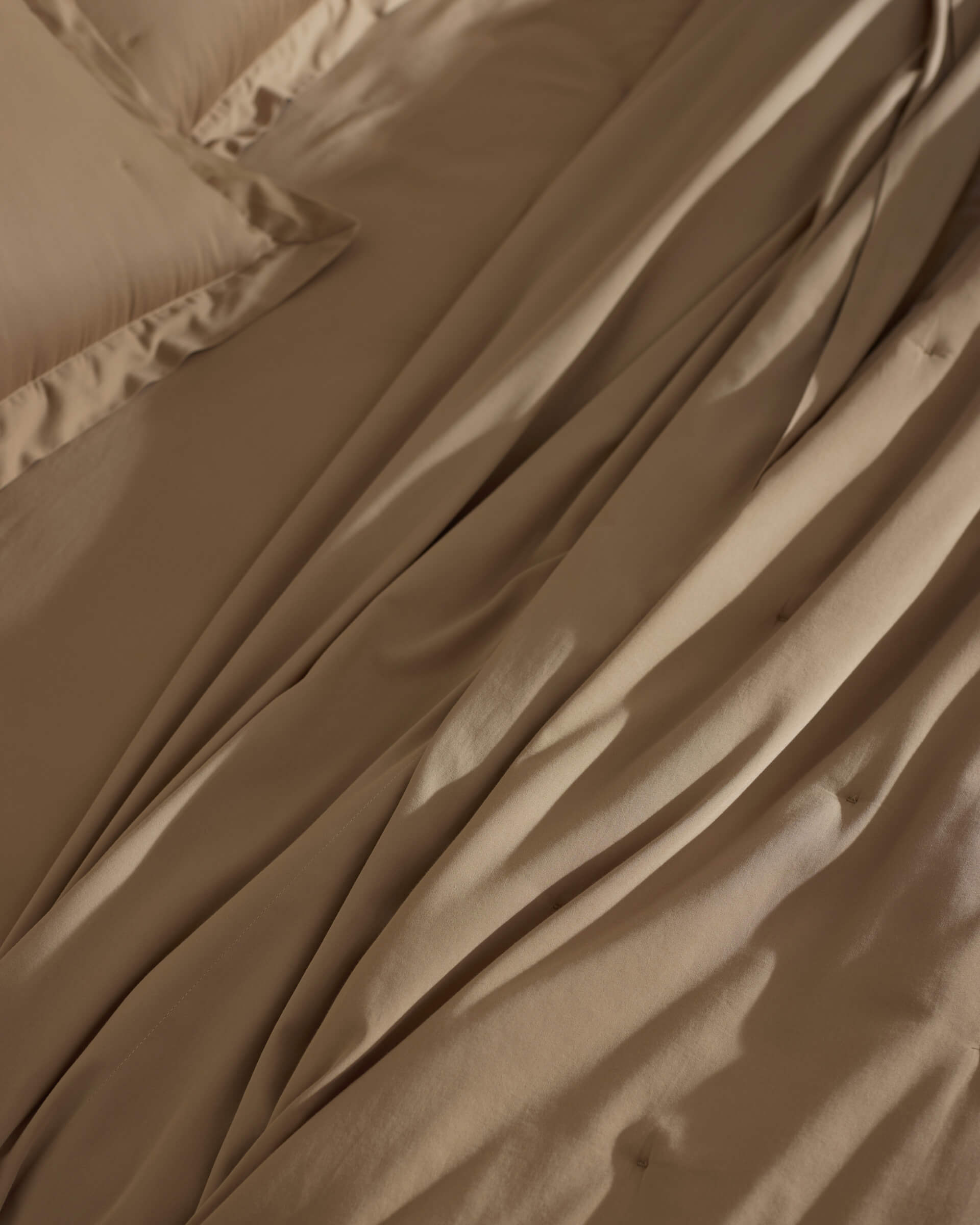 A close-up of a bed features the Cozy Earth Soft-Wash Cotton Quilt in tan, slightly wrinkled and naturally draped, showcasing its soft folds and gentle shadows.
