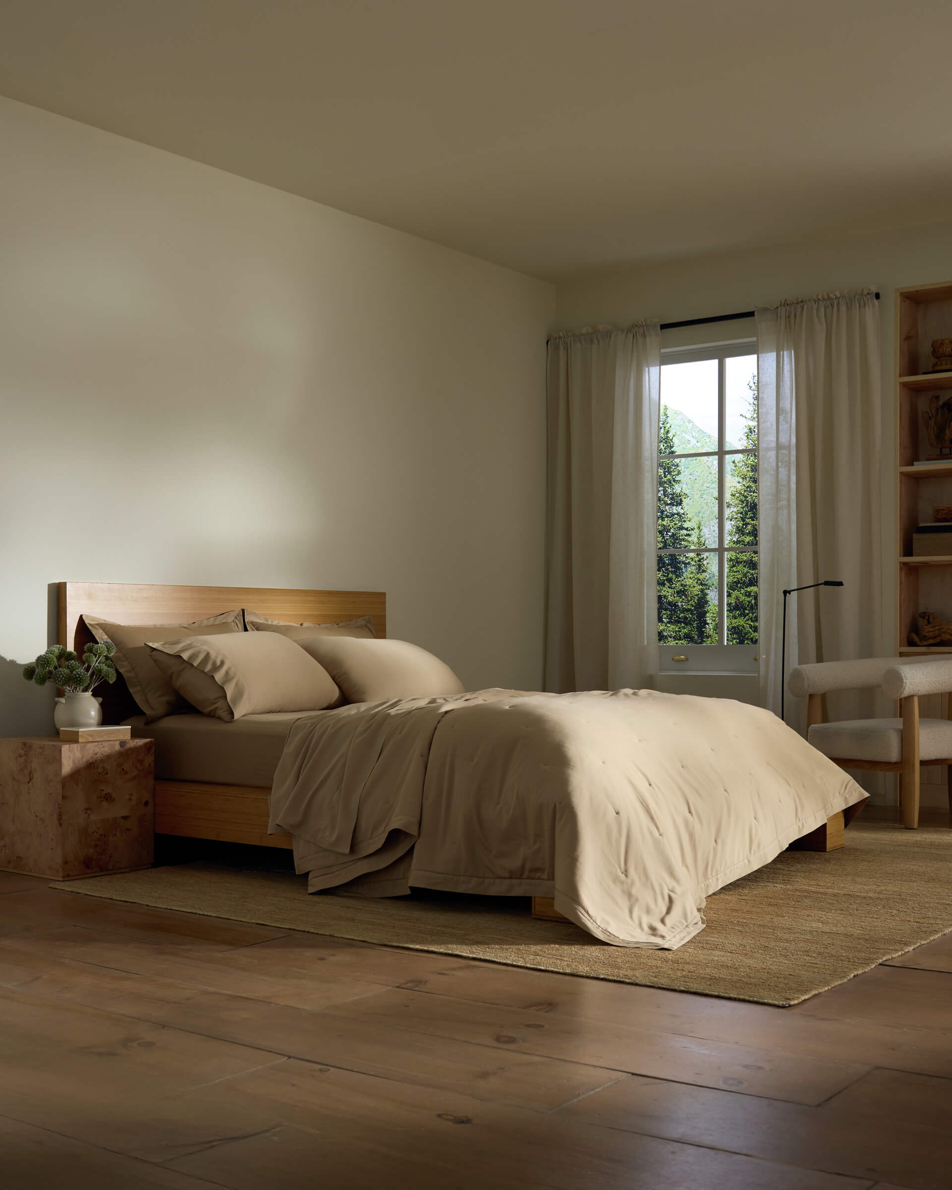 A cozy bedroom features the Cozy Earth Soft-Wash Cotton Quilt on a wooden bed, beige bedding, a bedside table with a plant, sheer-curtained window, cream armchair, wooden shelves, and a woven rug on a wooden floor.