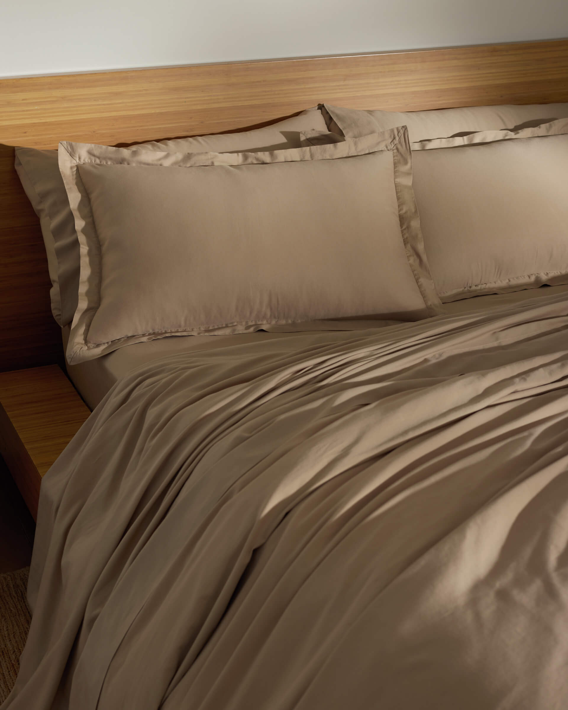 A softly lit room features a neatly made bed with the Cozy Earth Soft-Wash Cotton Duvet Set—light brown sheets, pillowcases, and matching comforter—set against a wooden headboard and side table.