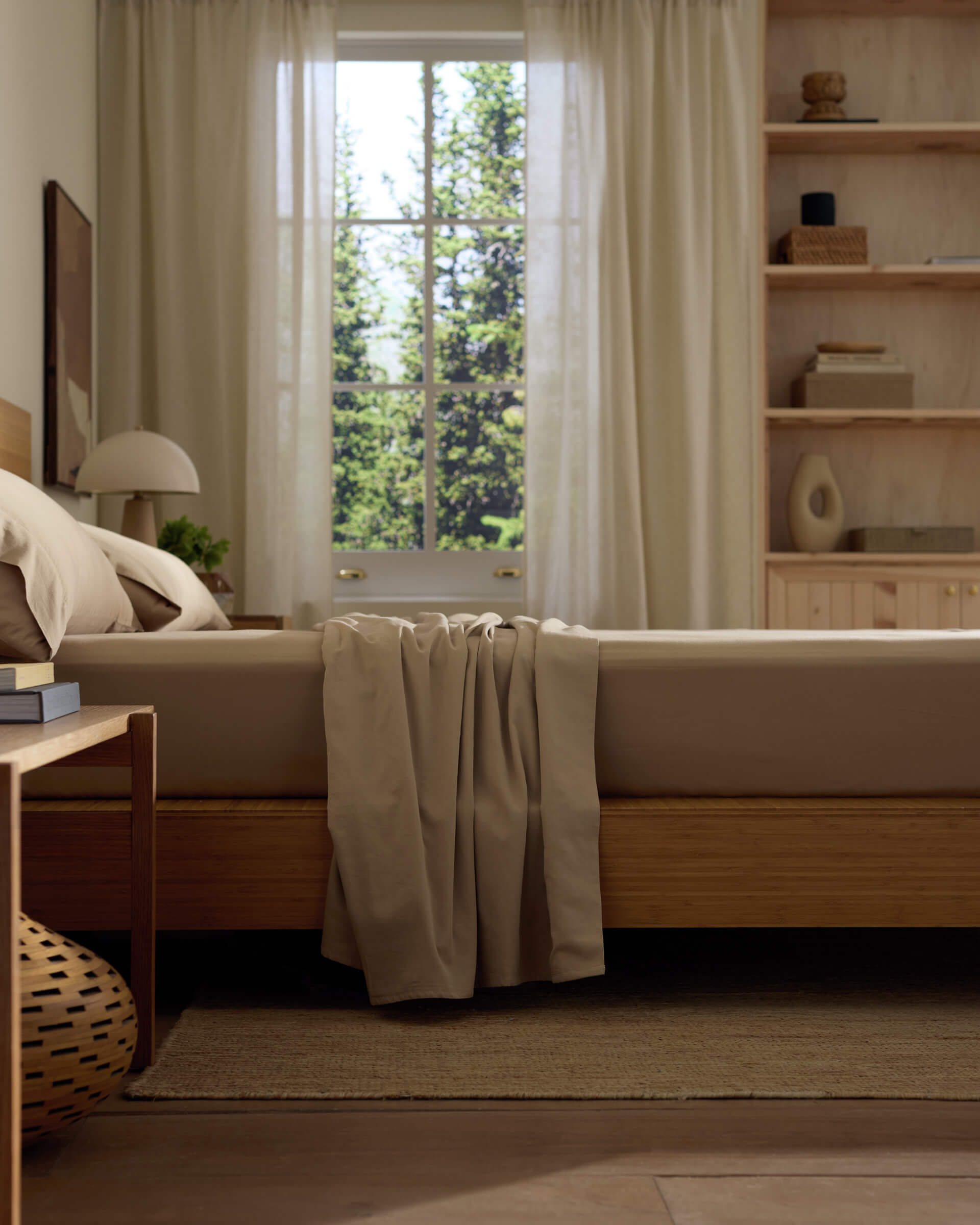 A sunlit bedroom features a neatly made bed with Cozy Earth's Soft-Wash Cotton Sheet Set, beige bedspread, wooden furniture, decor shelves, and a large window with sheer curtains framing green trees outside.