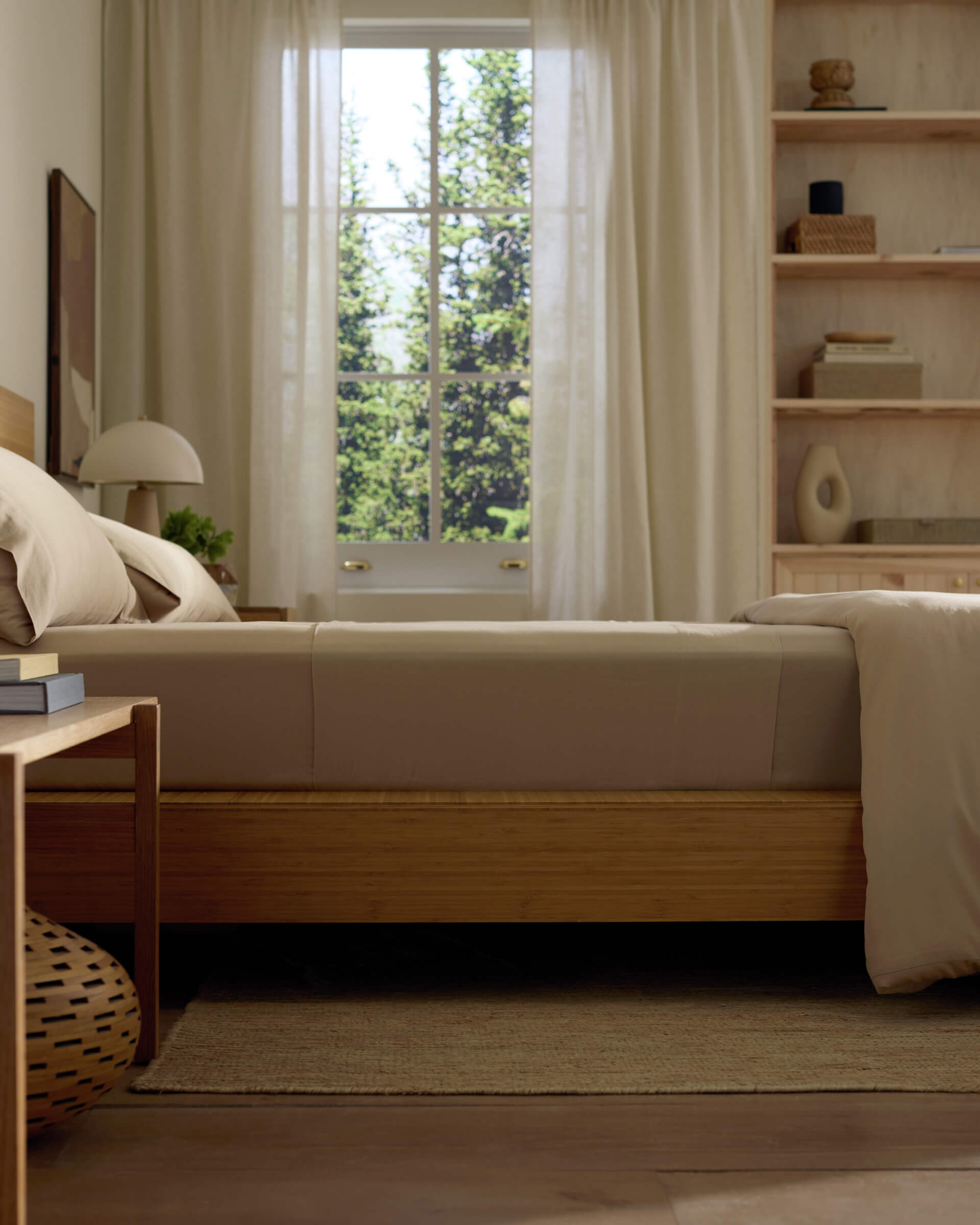 A serene bedroom features a Cozy Earth Soft-Wash Cotton Fitted Sheet on the bed, beige bedding, and white curtains. Sunlight streams in through the window, revealing green trees. Open shelving and a nightstand hold decor for a calm, minimalist vibe.