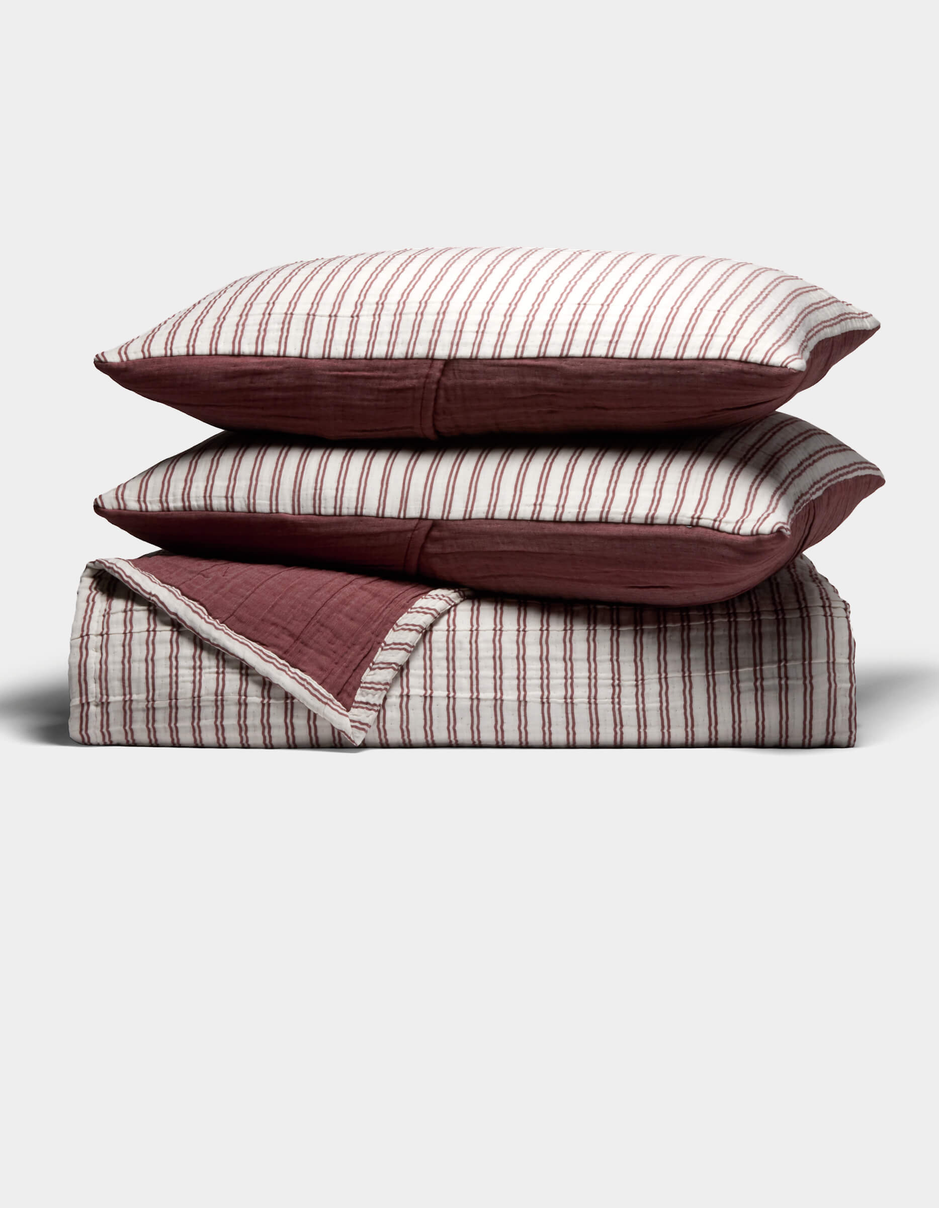 The Waterfront Coverlet Bundle by Cozy Earth features two pillows with red and white vertical stripes stacked on a matching folded quilt with a solid red underside, all displayed against a plain light background.