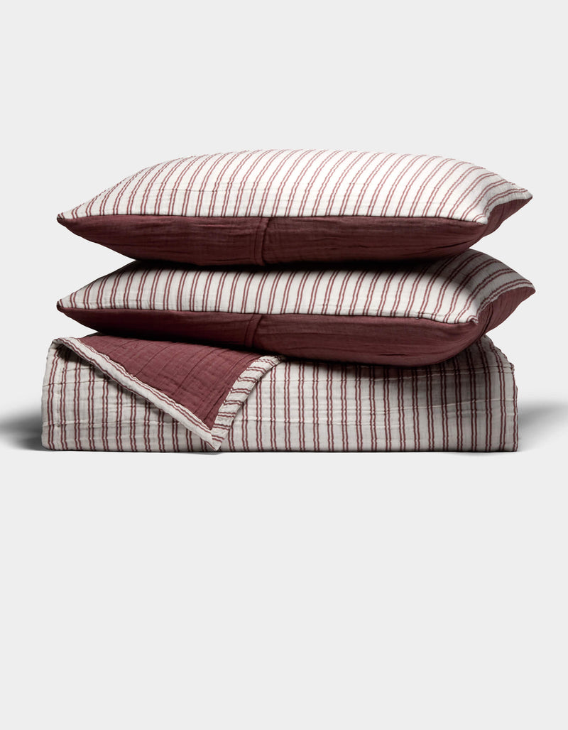 The Waterfront Coverlet Bundle by Cozy Earth features two pillows with red and white vertical stripes stacked on a matching folded quilt with a solid red underside, all displayed against a plain light background.