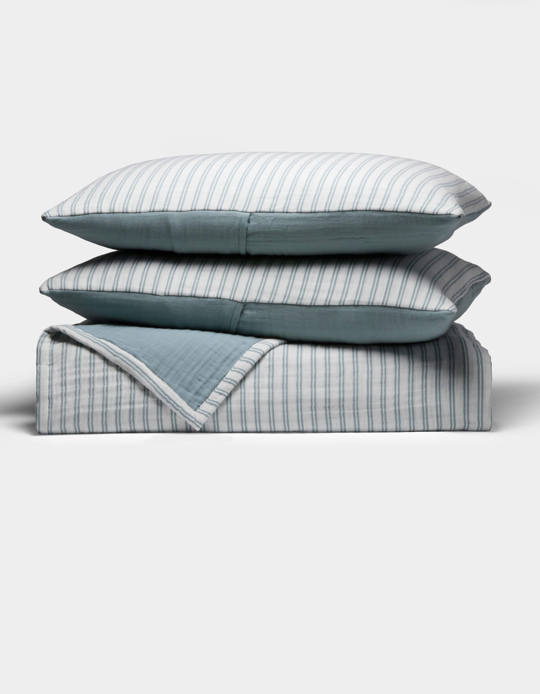 The Cozy Earth Waterfront Coverlet Bundle shows two pillows and a folded quilt stacked neatly, each with blue and white stripes on one side and solid blue on the other, all set against a plain white background.