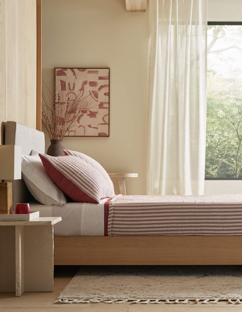 A modern bedroom features Cozy Earth's Waterfront Coverlet Bundle on a wooden bed, a beige rug, neutral walls, large windows with sheer curtains, and a vase with branches on the bedside table. Greenery can be seen outside.