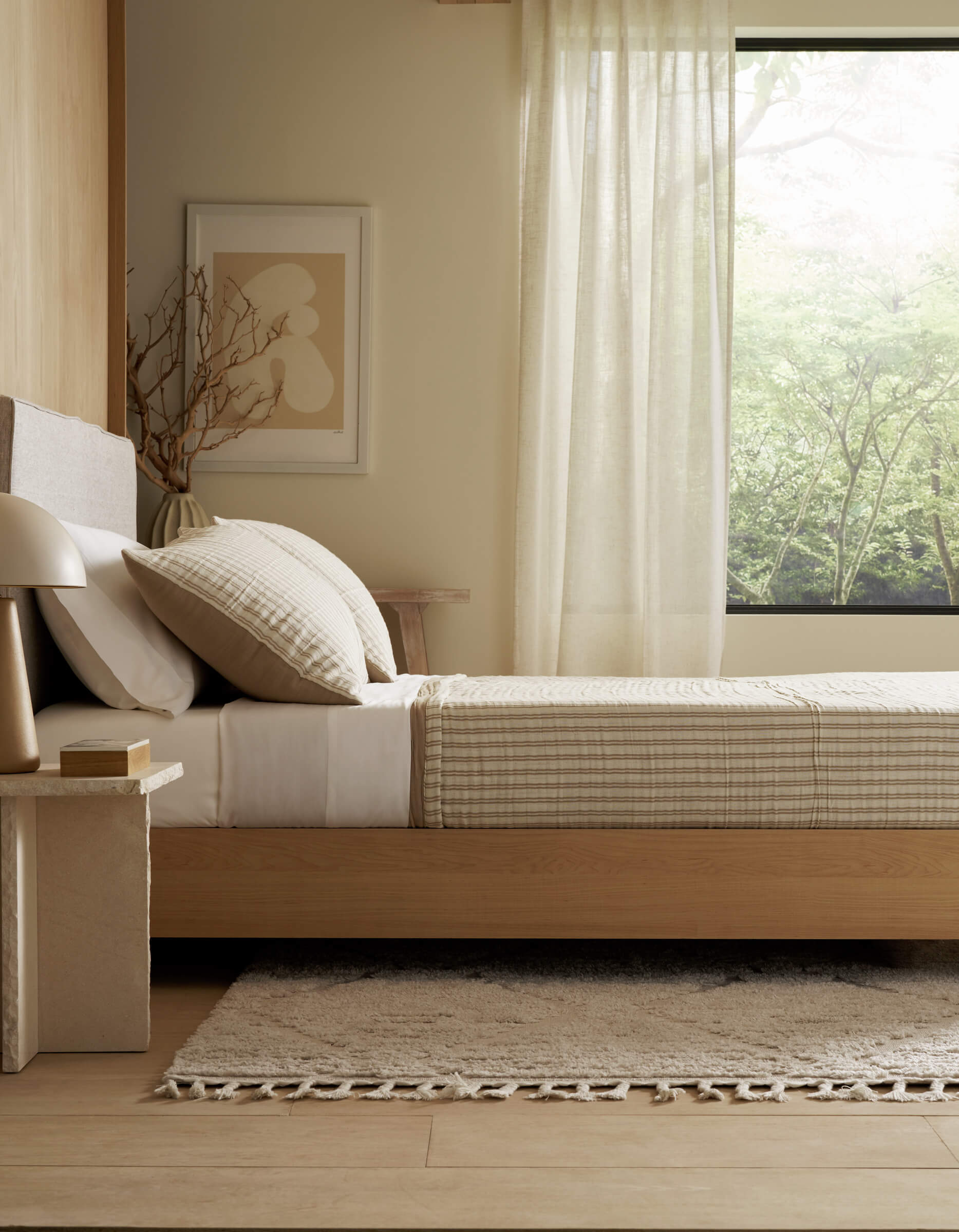 A minimal, cozy bedroom featuring Cozy Earth's Waterfront Coverlet, a light wooden bed, abstract wall art, a bedside table with lamp, and a large window with sheer curtains overlooking green trees.