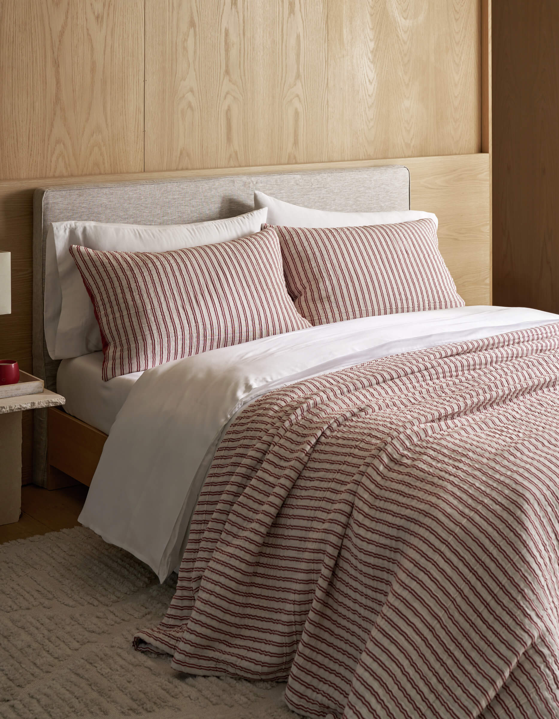 A neatly made bed features the Cozy Earth Waterfront Coverlet Bundle, white sheets, and a striped comforter with matching pillows, set against a light wood headboard and wall, complemented by a beige rug and a small bedside table.|Color:Currant Stripe