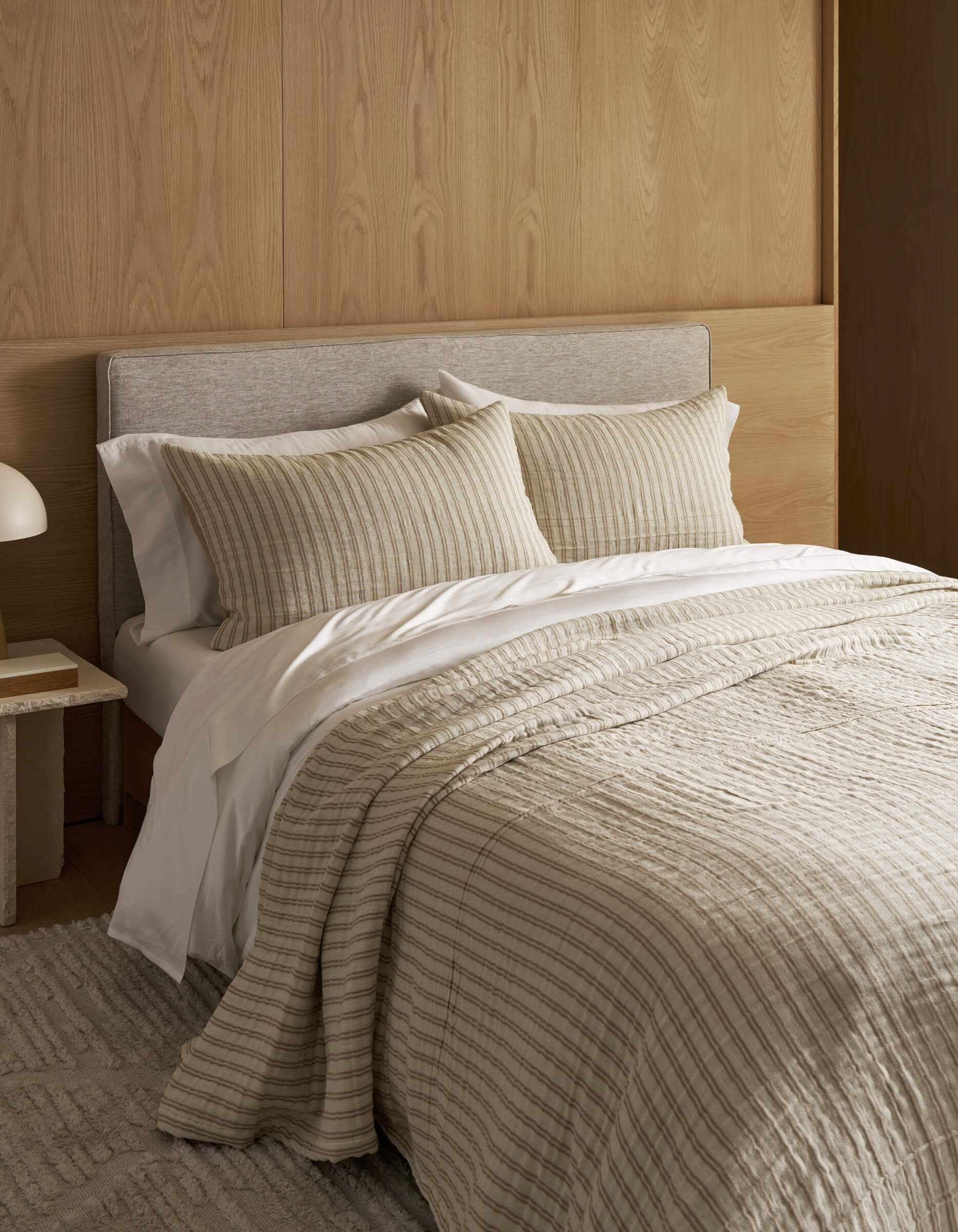 A neatly made bed features Cozy Earth's Waterfront Coverlet, two matching pillows, and a light gray upholstered headboard against a wooden wall. A small nightstand with a white lamp sits beside the bed.