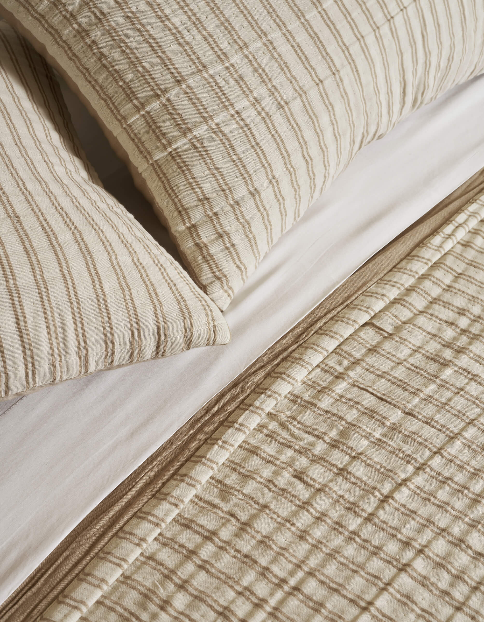 Close-up of a neatly made bed featuring Cozy Earth's Waterfront Coverlet, with cream and beige stripes on textured fabric and matching pillows, layered over crisp white sheets for a cozy look.