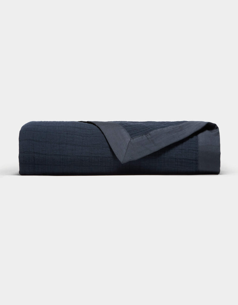 The Cozy Earth Waterfront Grid Matelasse Blanket is neatly folded in dark navy blue, showcasing its smooth, textured surface with one corner turned over, set against a plain light background.