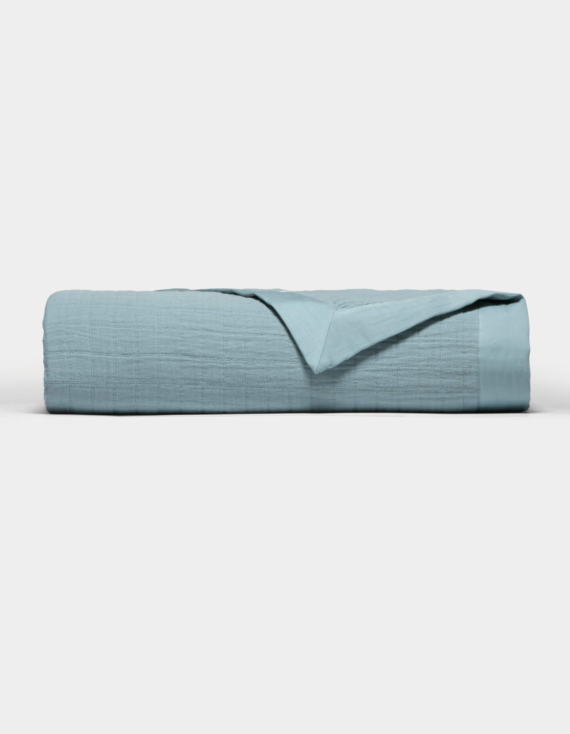 The Cozy Earth Waterfront Grid Matelasse Blanket in light blue is neatly folded against a plain white background, with the top layer turned back to show the inner fabric.
