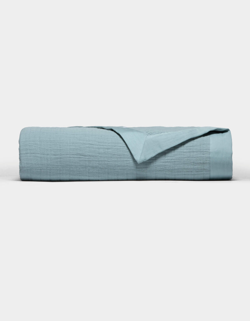 The Cozy Earth Waterfront Grid Matelasse Blanket in light blue is neatly folded against a plain white background, with the top layer turned back to show the inner fabric.