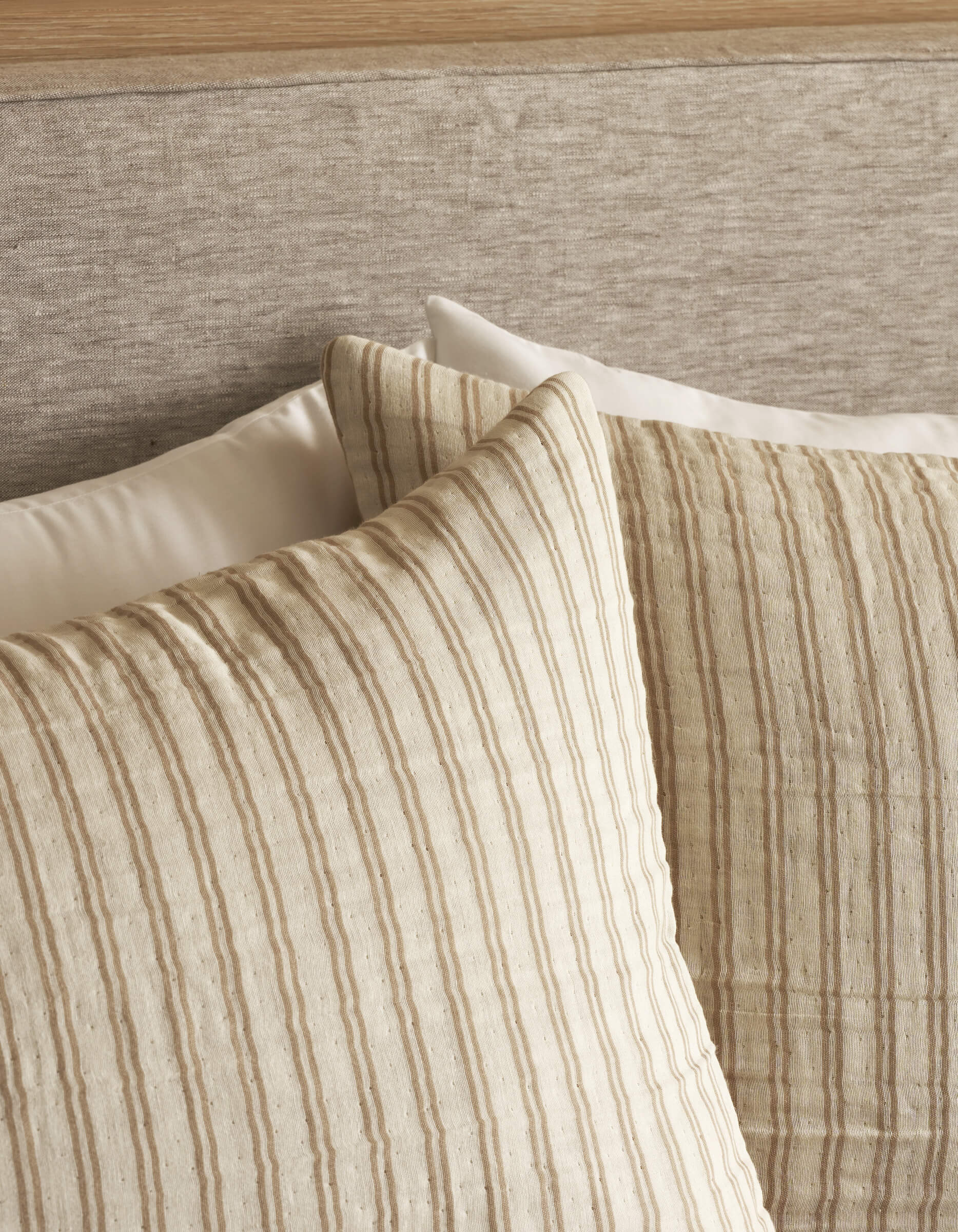 Two Waterfront Shams by Cozy Earth, cream-colored with thin vertical beige stripes, are propped against a beige fabric headboard, with a plain white pillow partially visible behind them for a calm, cozy look.
