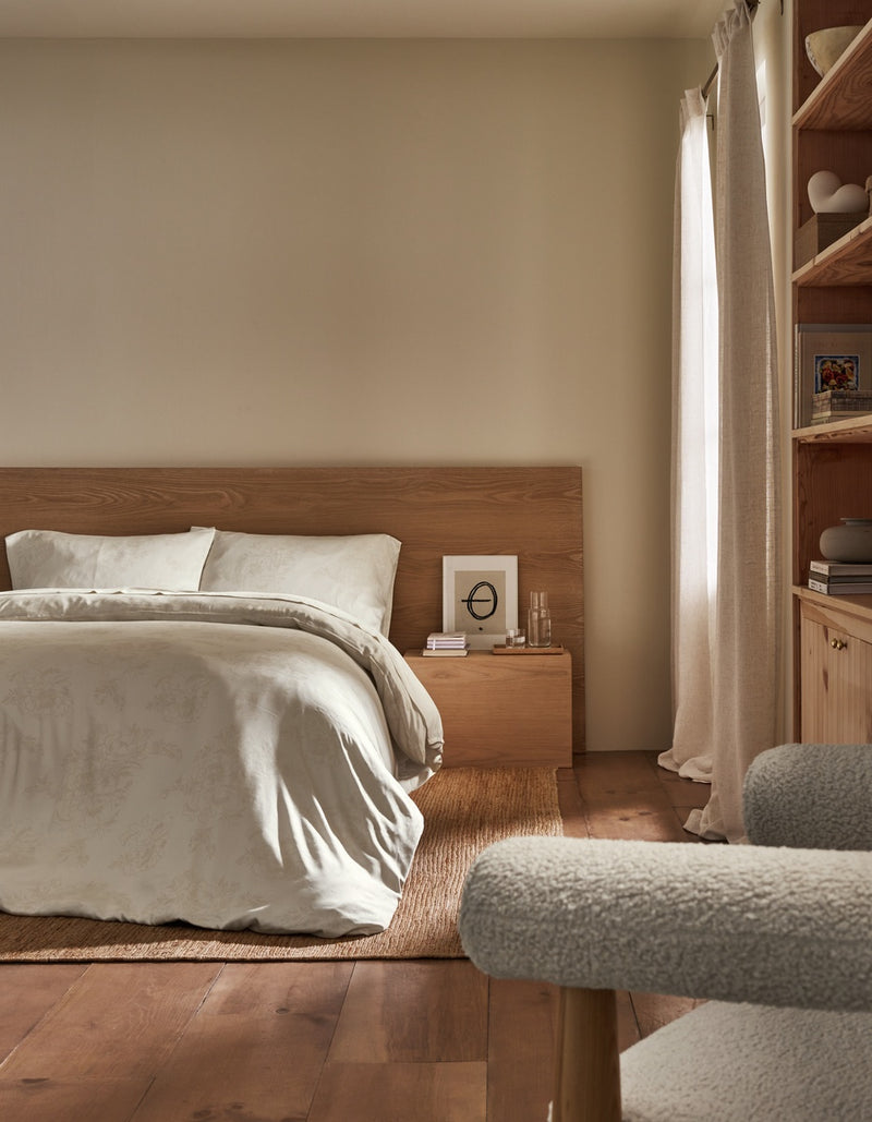 A minimalist bedroom scene featuring a bed with white bedding and a light wood headboard, styled in a warm neutral room with a wooden floor, beige walls, and a window with cream curtains.