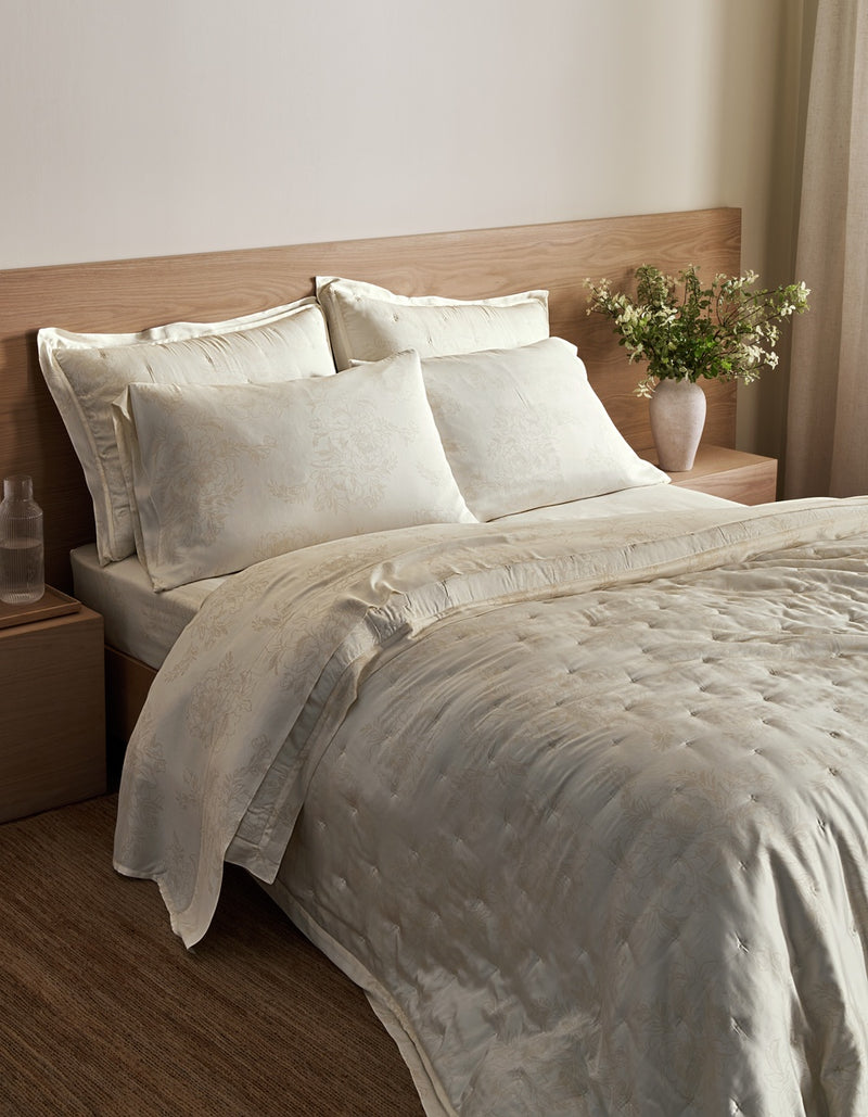 A neatly made bed in a bedroom setting, dressed with a textured cream quilt and matching pillow shams, set against a light wood headboard and bedside tables.