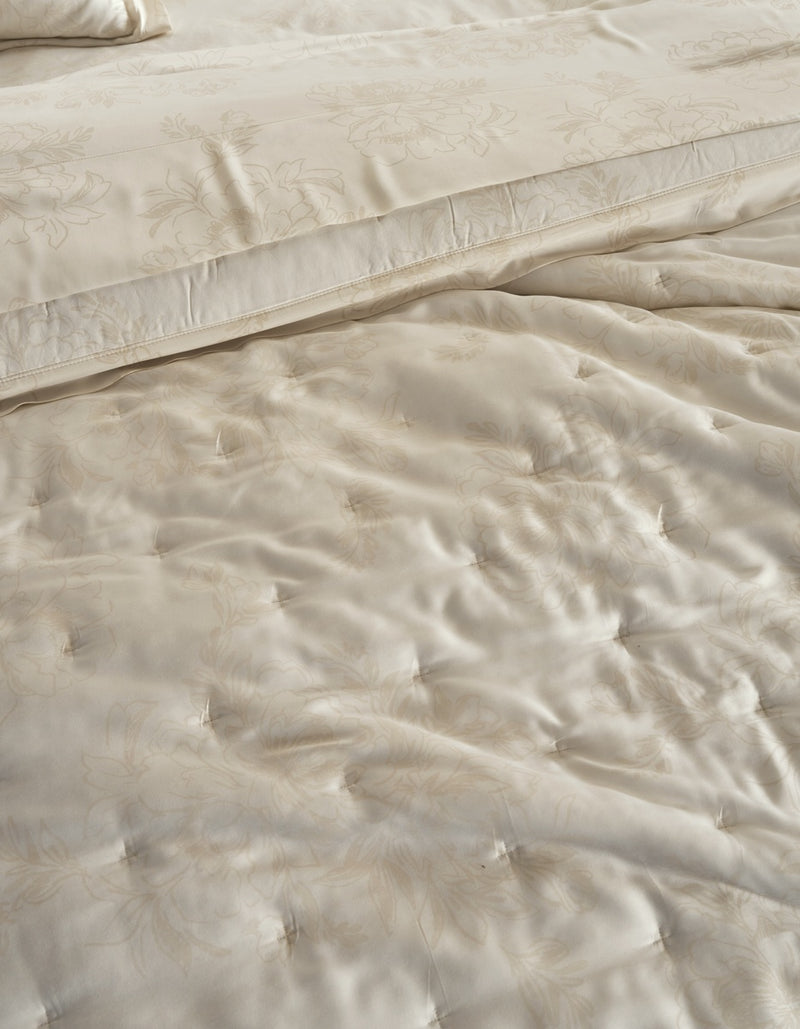 Close-up of a cream-colored quilted bedding set with a subtle floral jacquard pattern, showing the texture of the tufted comforter and matching pillowcase.