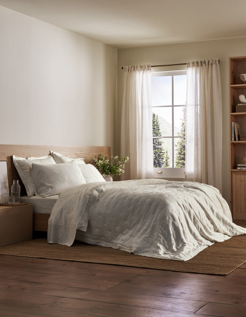 A rustic, minimalist bedroom featuring a bed dressed in white and beige linen bedding, set against a light wood headboard and footboard. The room has warm beige walls, sheer curtains by a bright window, a wooden bookshelf, and dark wood flooring.