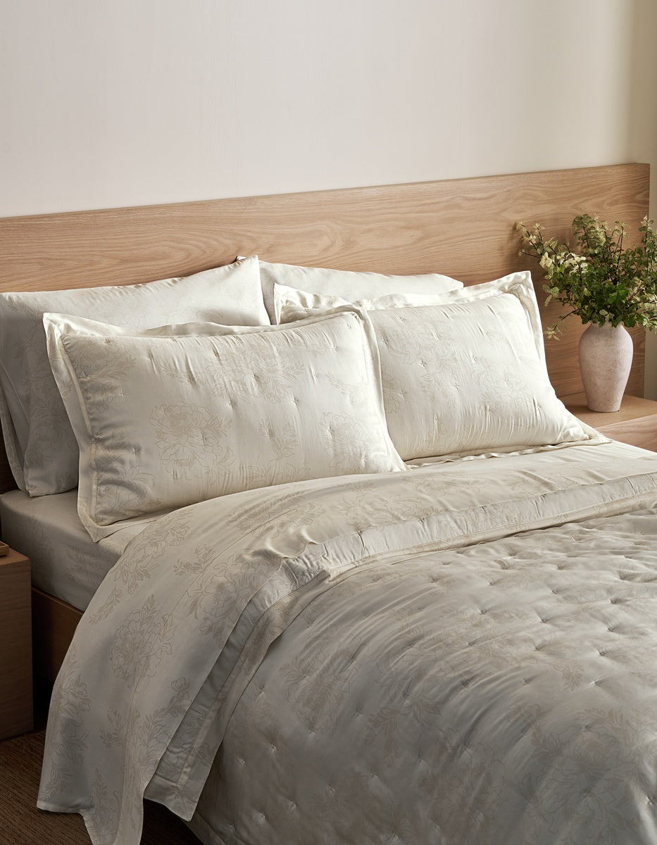 A neatly made bed with textured cream-colored bedding, including a quilted duvet cover and matching pillow shams, set against a light wood headboard and a neutral wall.