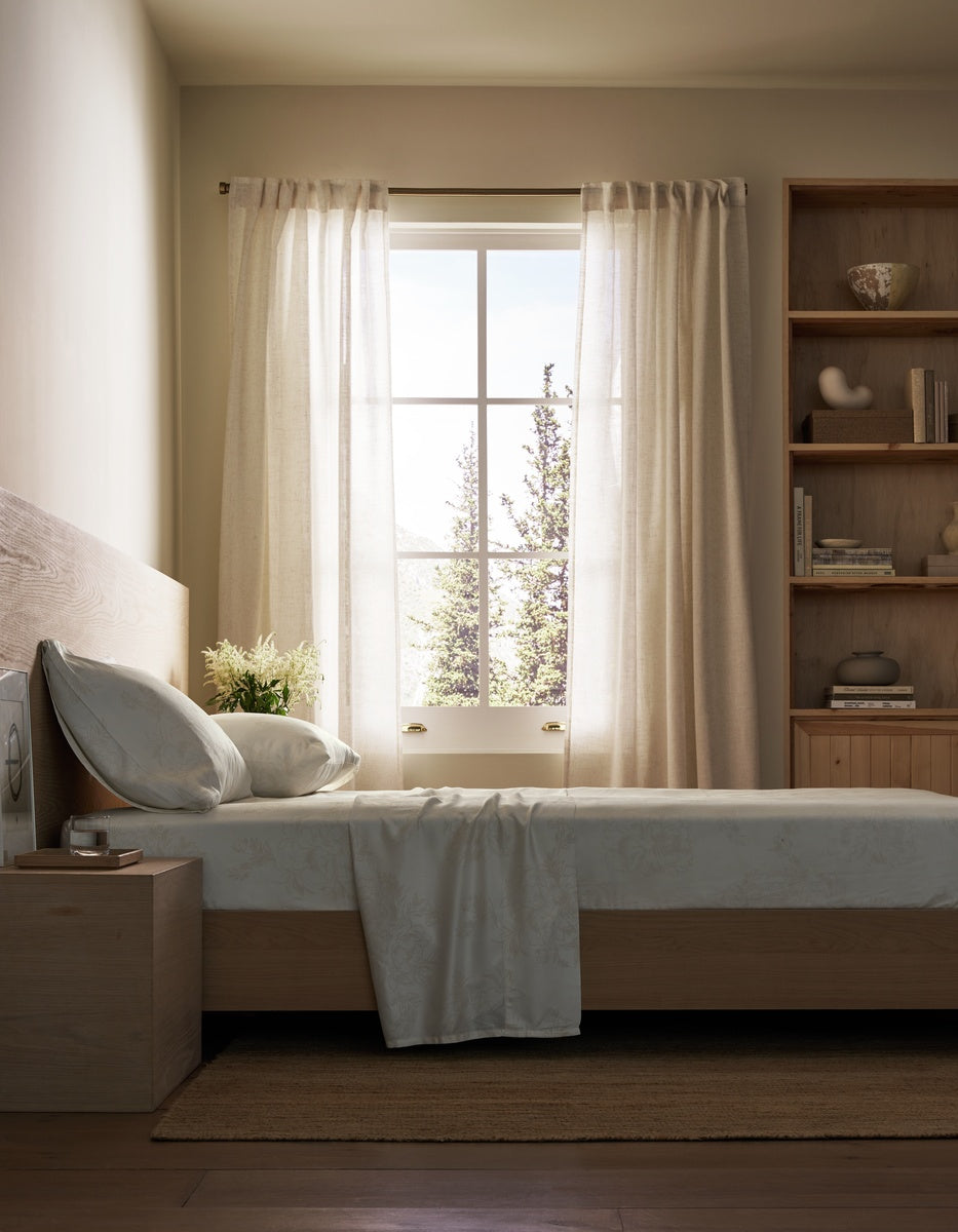 A minimalist bedroom scene featuring a light wood bed frame with white bedding, sheer cream curtains, a wooden headboard, and a matching wooden shelving unit against a warm beige wall. 