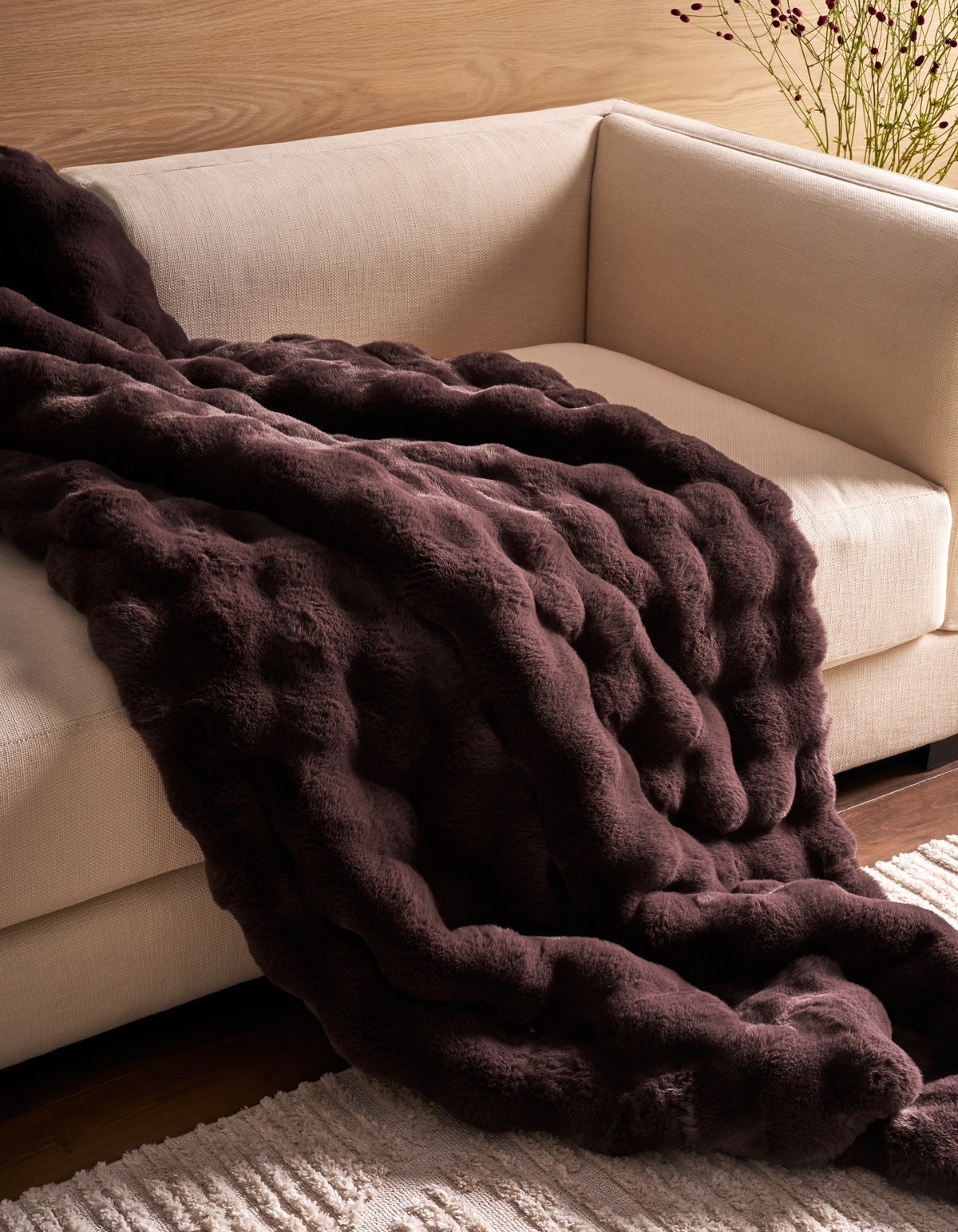 The Cozy Earth Bubble Cuddle Blanket rests atop a light beige sofa, complemented by a cream-colored rug and a wooden wall in the background.