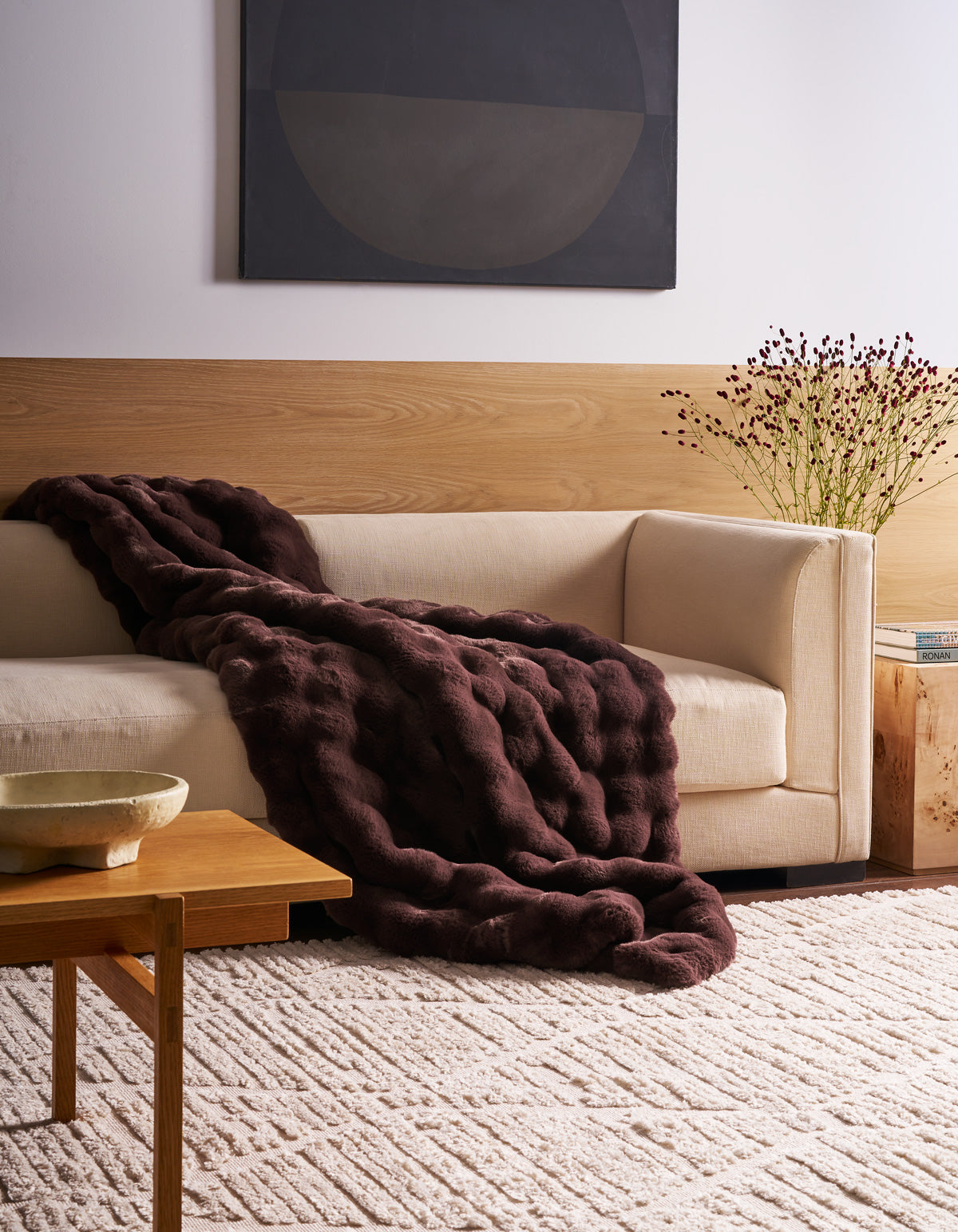 A modern living room featuring a cream-colored sofa, adorned with the Cozy Earth Bubble Cuddle Blanket, complemented by a dark purple textured blanket, a wooden table, a patterned rug, and minimalist decor.