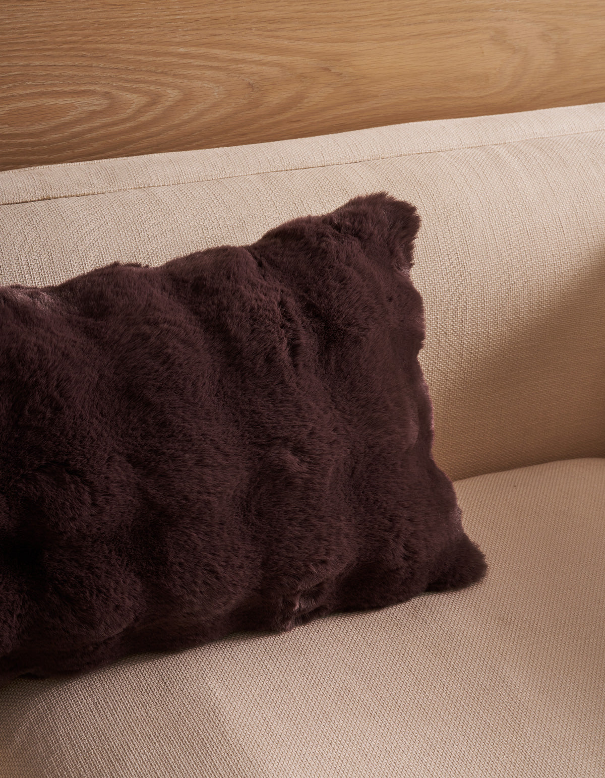 A Bubble Cuddle Pillow by Cozy Earth in a rich dark brown hue rests on a light beige textured sofa, with a wooden wall panel visible in the background.