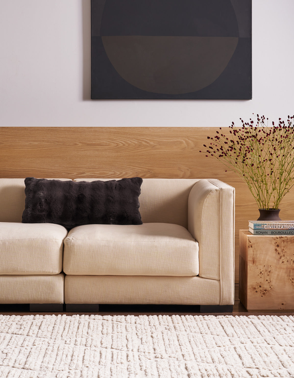 A beige sofa with a Cozy Earth Bubble Cuddle Pillow sits beside a wooden side table holding books and a vase with twigs. A textured white rug covers the floor, and abstract art hangs on the wall above wood paneling.