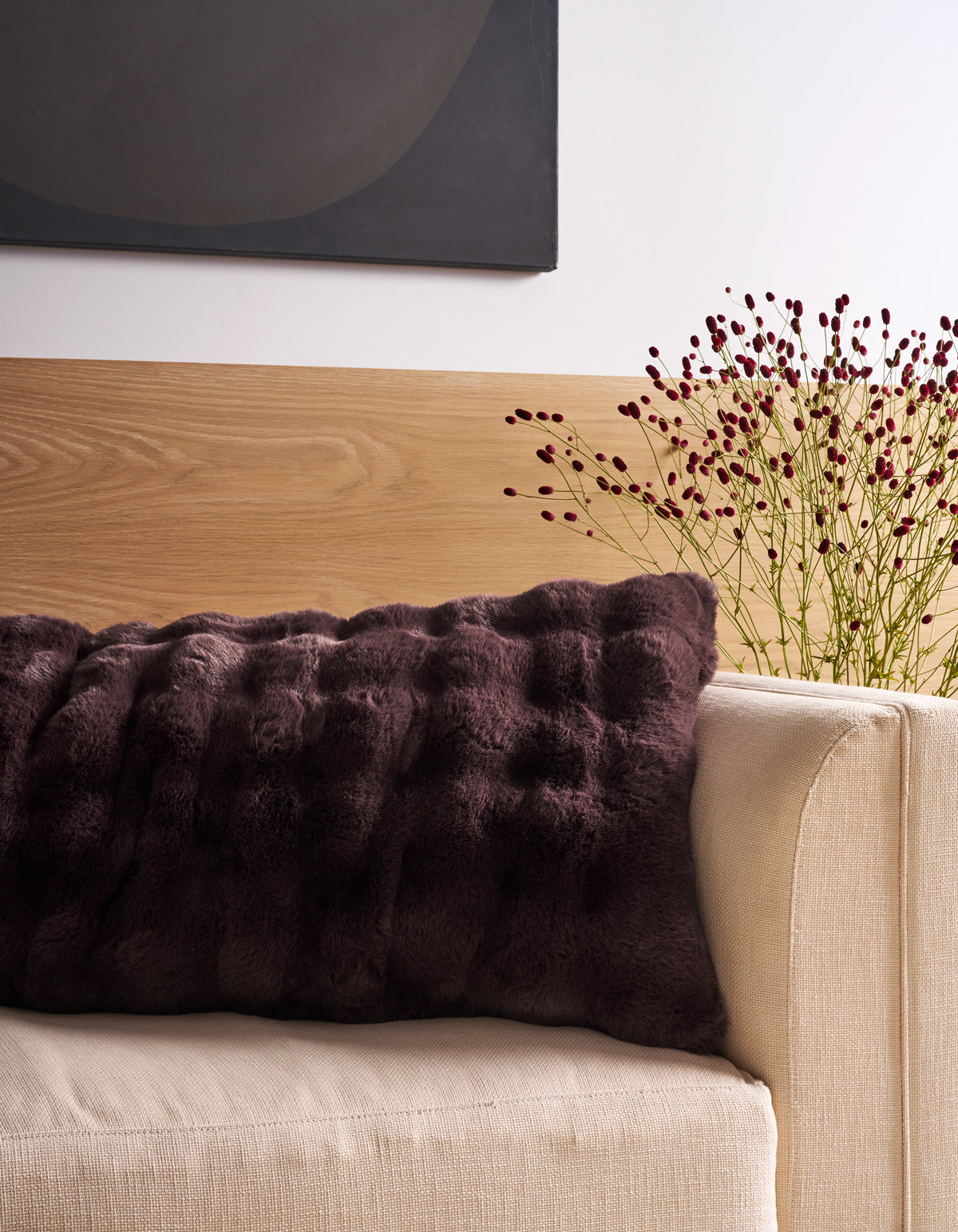A cream sofa adorned with the Cozy Earth Bubble Cuddle Pillow—a large, dark brown textured accent—sits below a modern black-and-wood wall art piece, while delicate sprigs of small red flowers in a vase rest beside the sofa.
