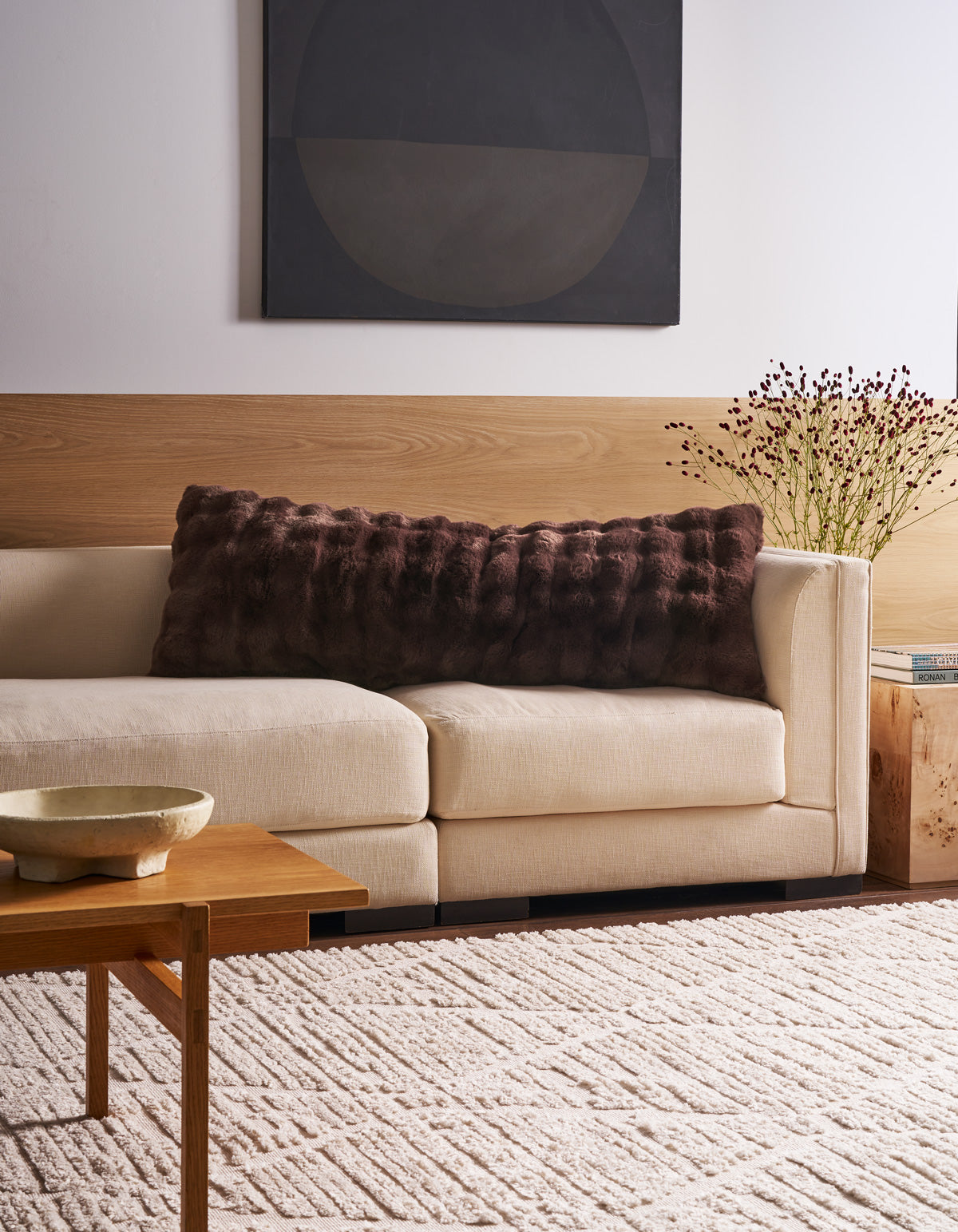 A modern living room featuring a cream sofa adorned with the Bubble Cuddle Pillow by Cozy Earth, abstract wall art, a wooden coffee table with a decorative bowl, a side table topped with books and dried flowers, and a patterned beige rug.