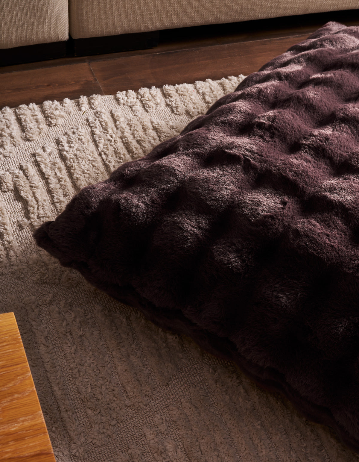 A Cozy Earth Bubble Cuddle Pillow in deep purple, featuring a textured design, rests on a light, patterned carpet. In the background, part of a tan sofa and a wooden table can be seen.