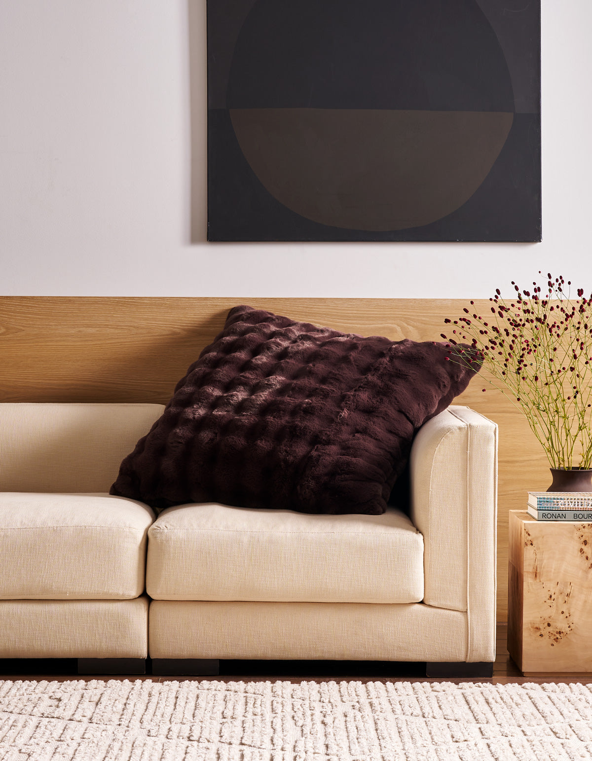 A cream-colored sofa with the large, dark-textured Bubble Cuddle Pillow from Cozy Earth sits against a wood-paneled wall. A black abstract painting hangs above, and a wooden side table with a vase of dried flowers is nearby.