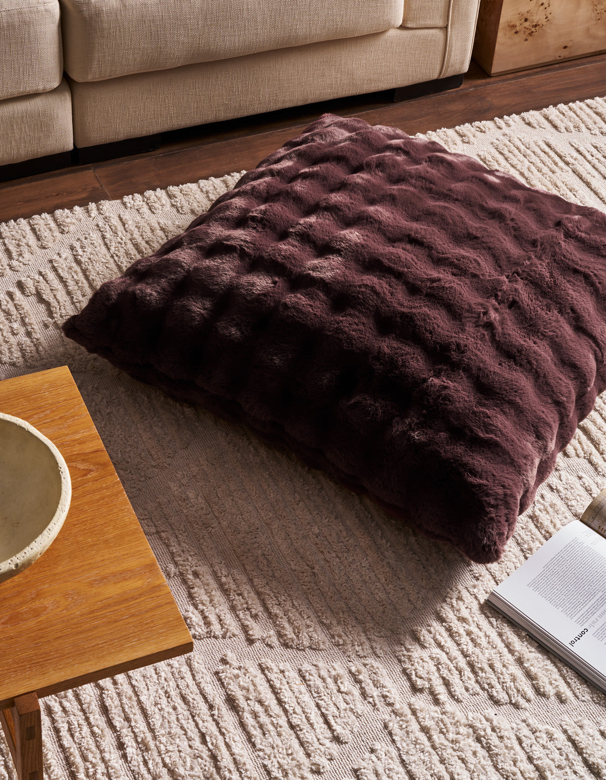 A spacious, textured Bubble Cuddle Pillow by Cozy Earth in dark brown rests atop a cream patterned rug beside a wooden coffee table and an open magazine, creating a cozy living room atmosphere.