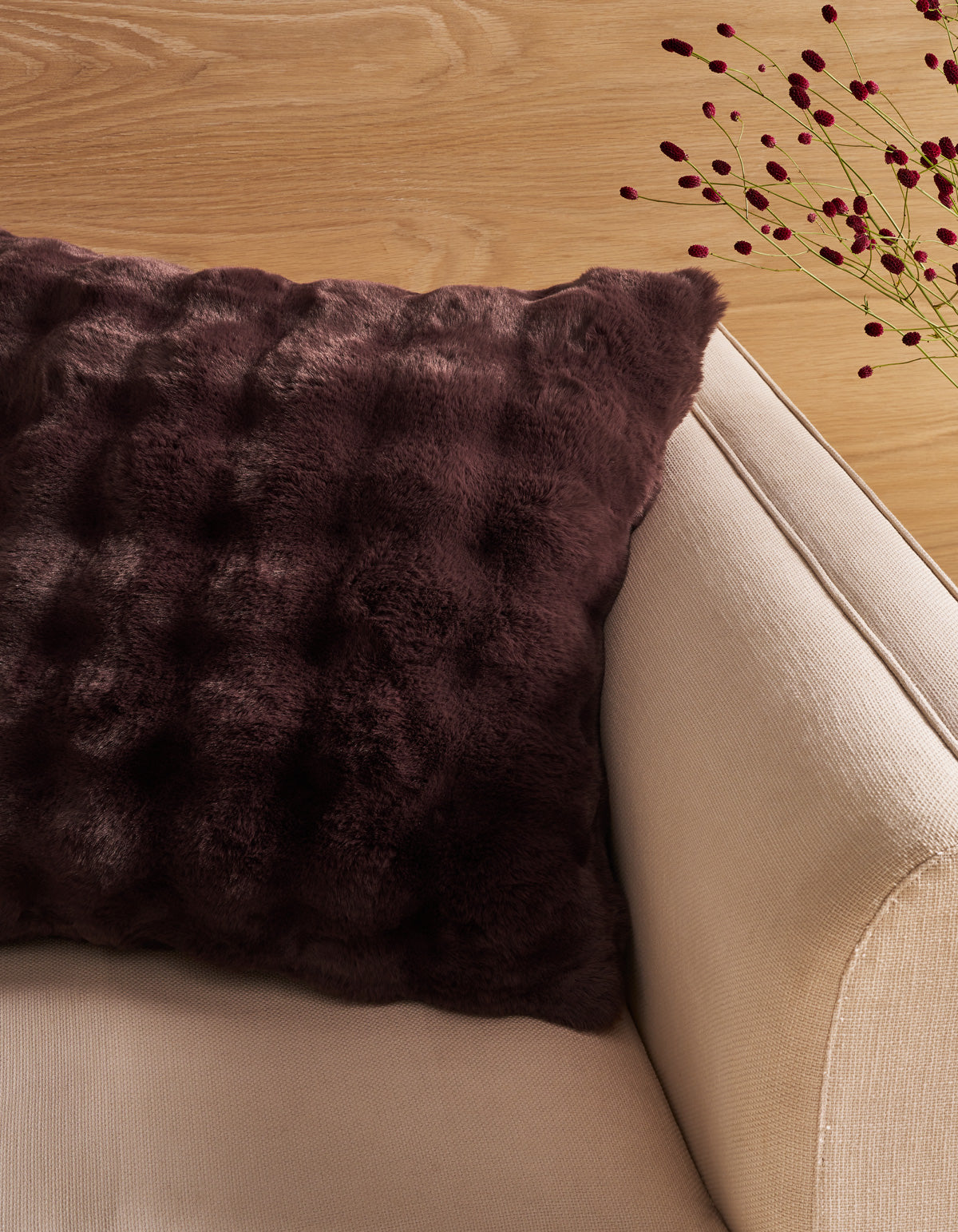 A Bubble Cuddle Pillow by Cozy Earth, featuring a dark brown textured faux fur design, rests on a beige armchair beside a light wooden surface with delicate red berry stems in the background.