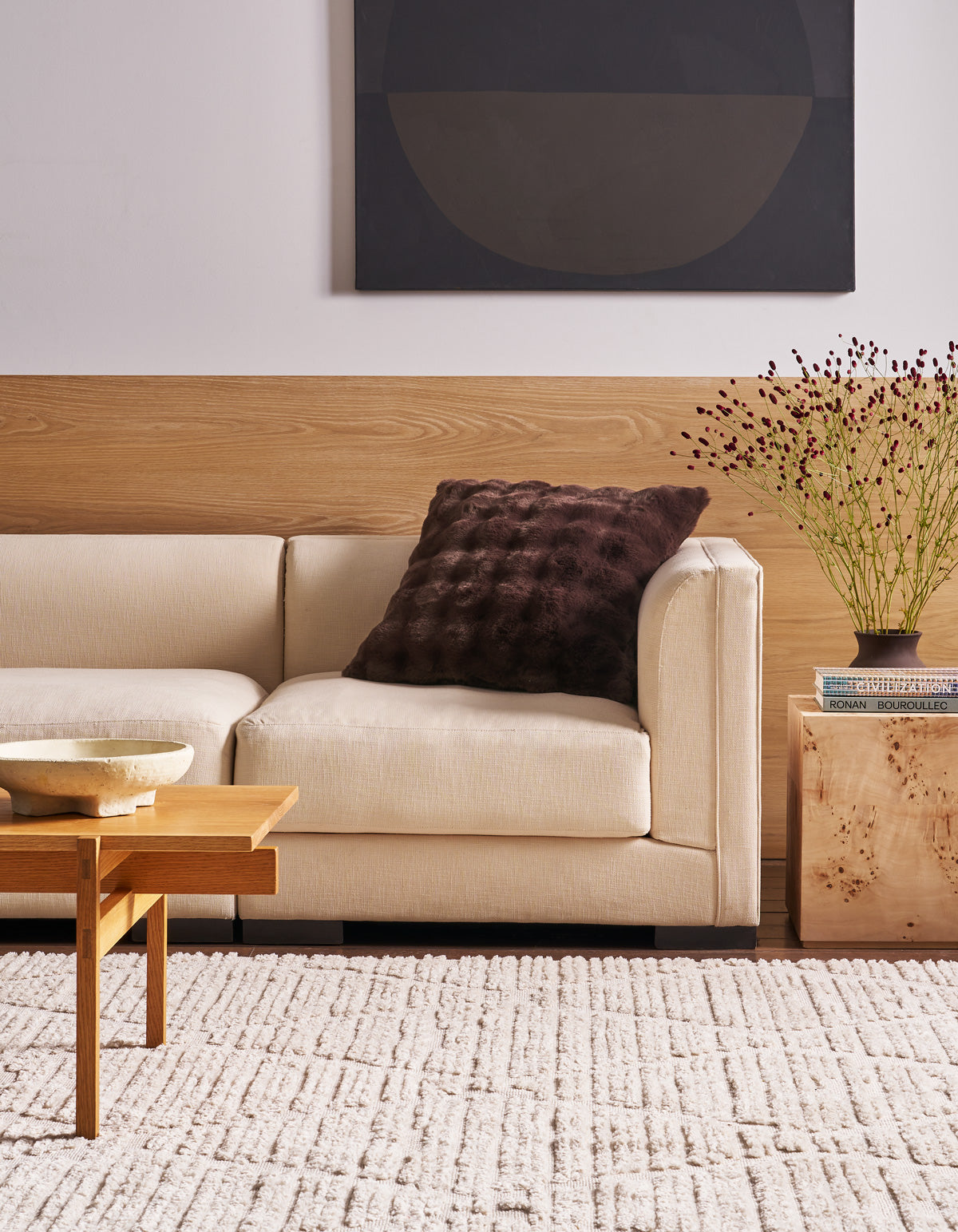 A modern living room with a beige sofa, a Cozy Earth Bubble Cuddle Pillow in a dark textured finish, a wooden coffee table, light rug, side table with books and a vase of flowers, and abstract art on the wall.