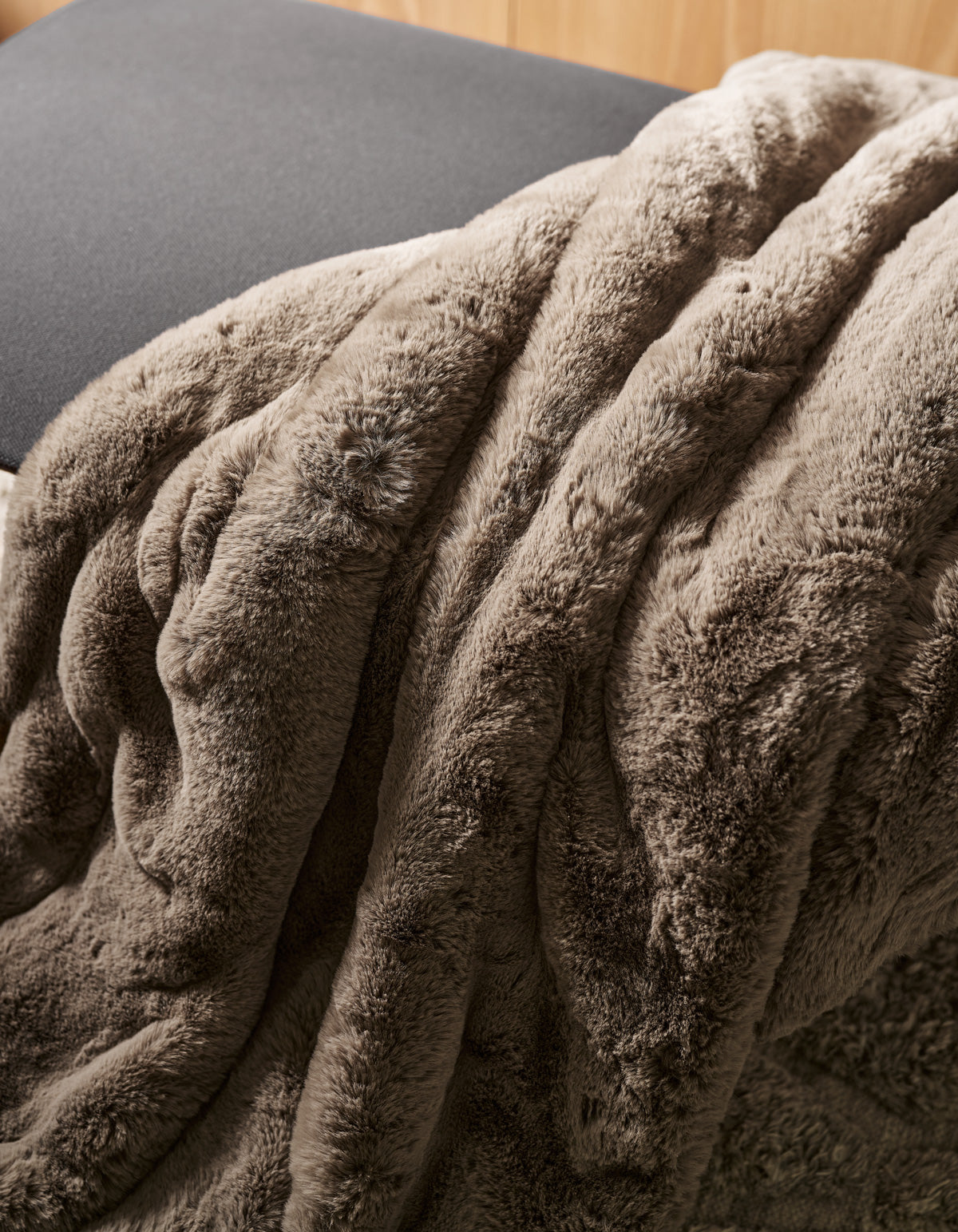 The Channel Cuddle Blanket by Cozy Earth, in a soft brown plush fabric, is draped over the edge of a gray cushion or chair, creating a cozy and inviting atmosphere.