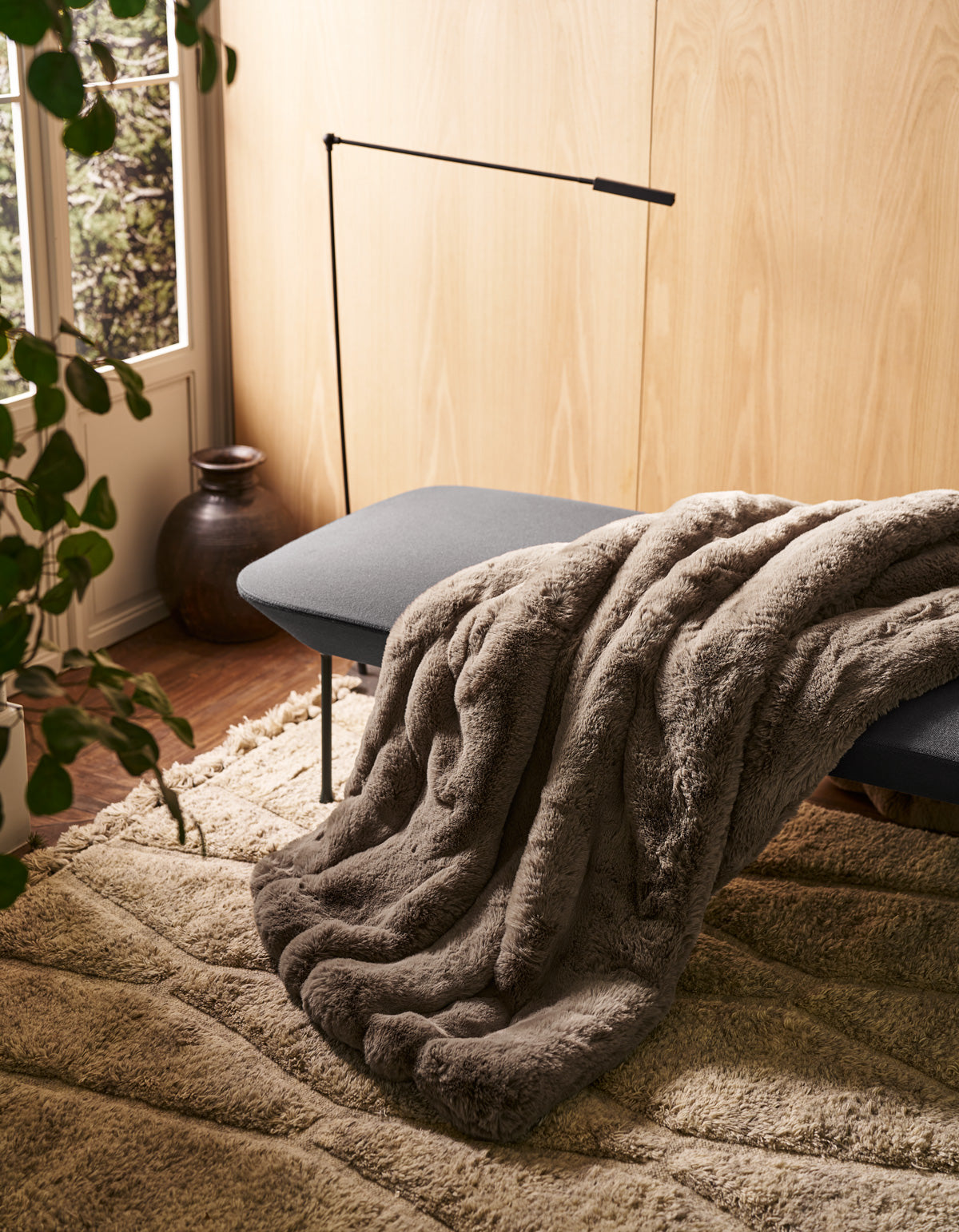 The Channel Cuddle Blanket from Cozy Earth, in a soft gray hue, is draped over a cushioned bench near a window. Nearby, a plant and a ceramic vase add natural charm as sunlight streams in, illuminating the textured beige rug and highlighting the warmth of the wooden wall.
