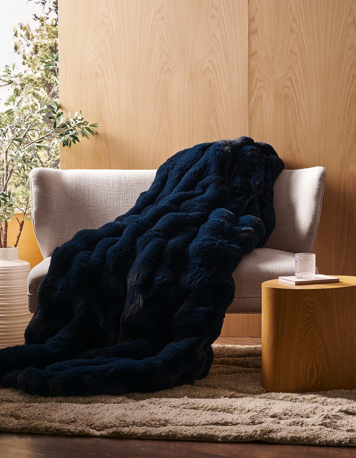 A Cozy Earth Double Bubble Cuddle Blanket in dark blue is draped over a light gray sofa, beside a round wooden side table holding a glass of water, all set in a cozy room with wood-paneled walls and a beige rug.