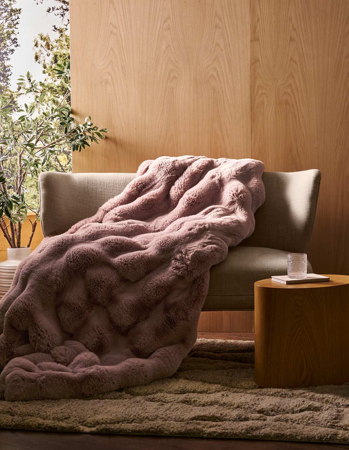 A beige sofa with the Cozy Earth Double Bubble Cuddle Blanket in textured pink draped over it sits beside a small wooden table holding a glass of water and a book, set in a cozy room with wood-paneled walls and a potted plant.