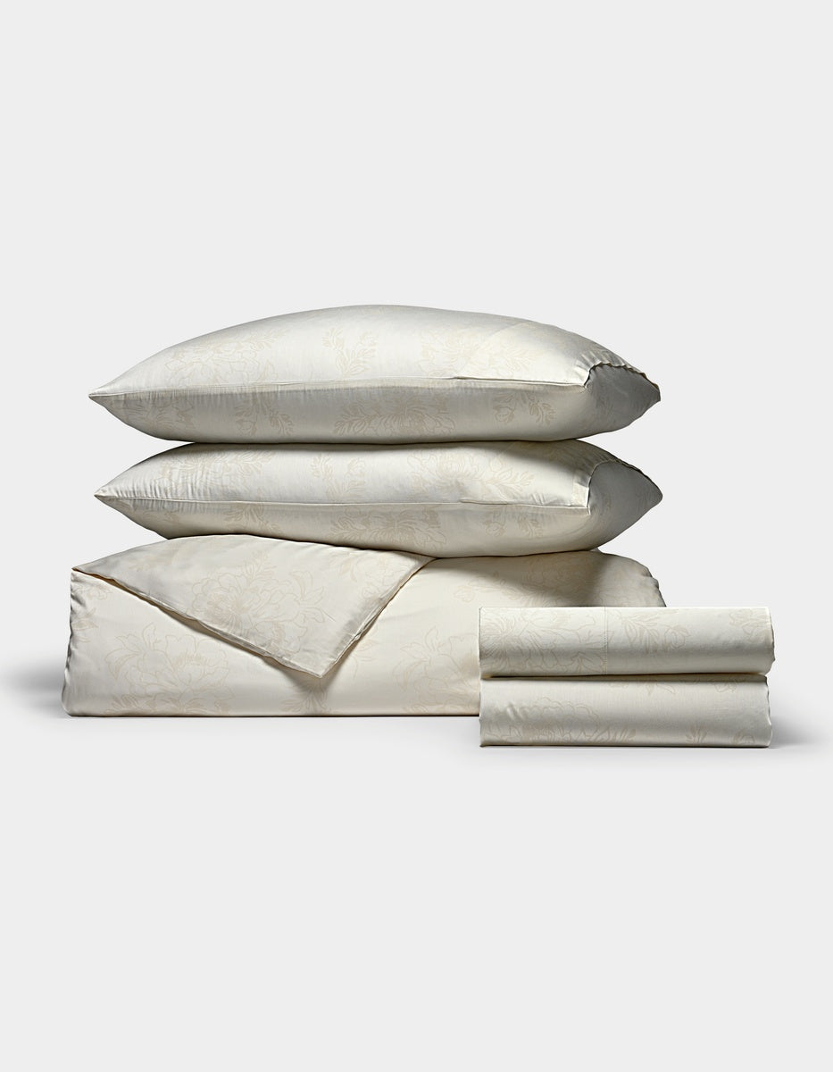 A Cozy Earth Bamboo Bedding Deluxe Bundle in cream, featuring a subtle floral pattern, with two pillows stacked atop a folded duvet and neatly folded sheets, displayed on a plain white background.|Color:Sonoma Floral in Vintage White