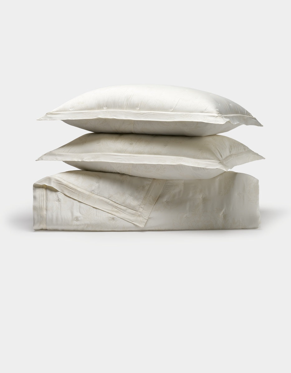 Two white Cozy Earth pillows are stacked atop a folded Bamboo Quilt Bundle, all crafted from a shiny, smooth fabric and set against a plain light background.|Color:Sonoma Floral in Vintage White