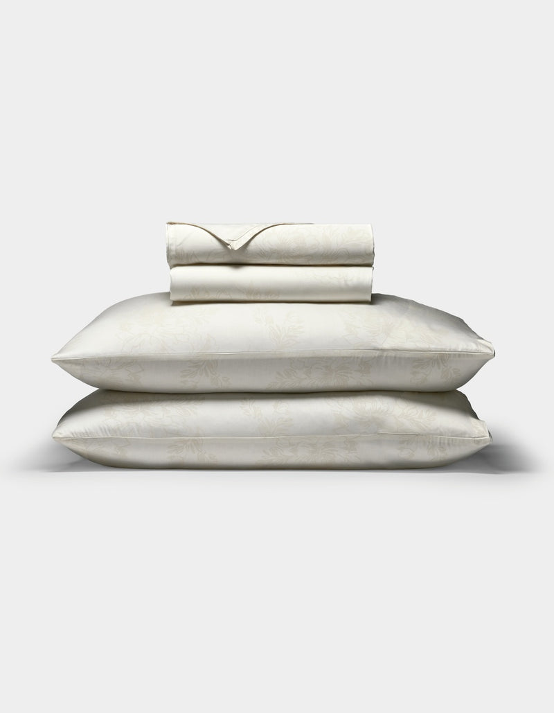 A Cozy Earth Bamboo Sheet Set in cream, featuring a subtle floral pattern, with two pillows in matching pillowcases stacked beneath neatly folded sheets, displayed on a plain white background.
