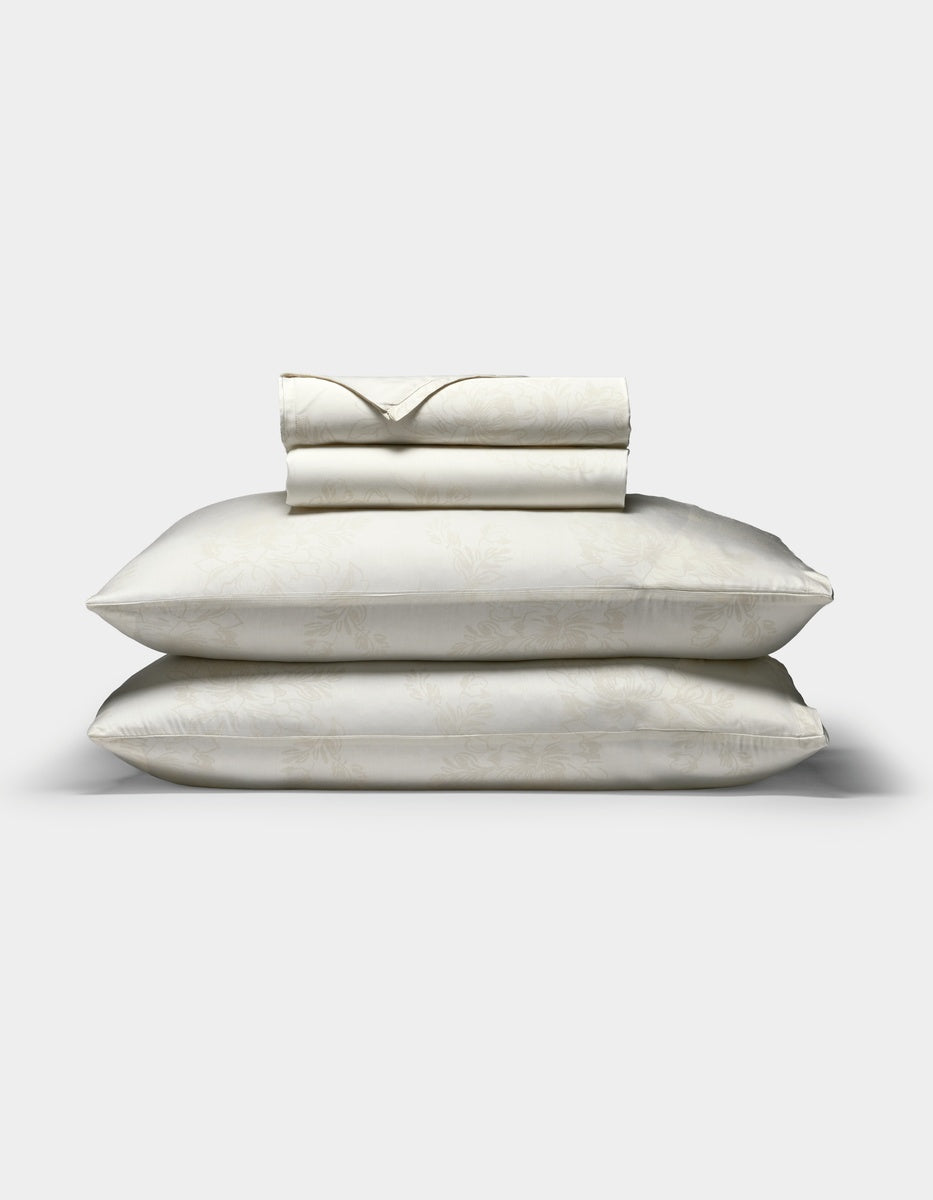 A Cozy Earth Bamboo Sheet Set in cream, featuring a subtle floral pattern, with two pillows in matching pillowcases stacked beneath neatly folded sheets, displayed on a plain white background.|Color:Sonoma Floral in Vintage White