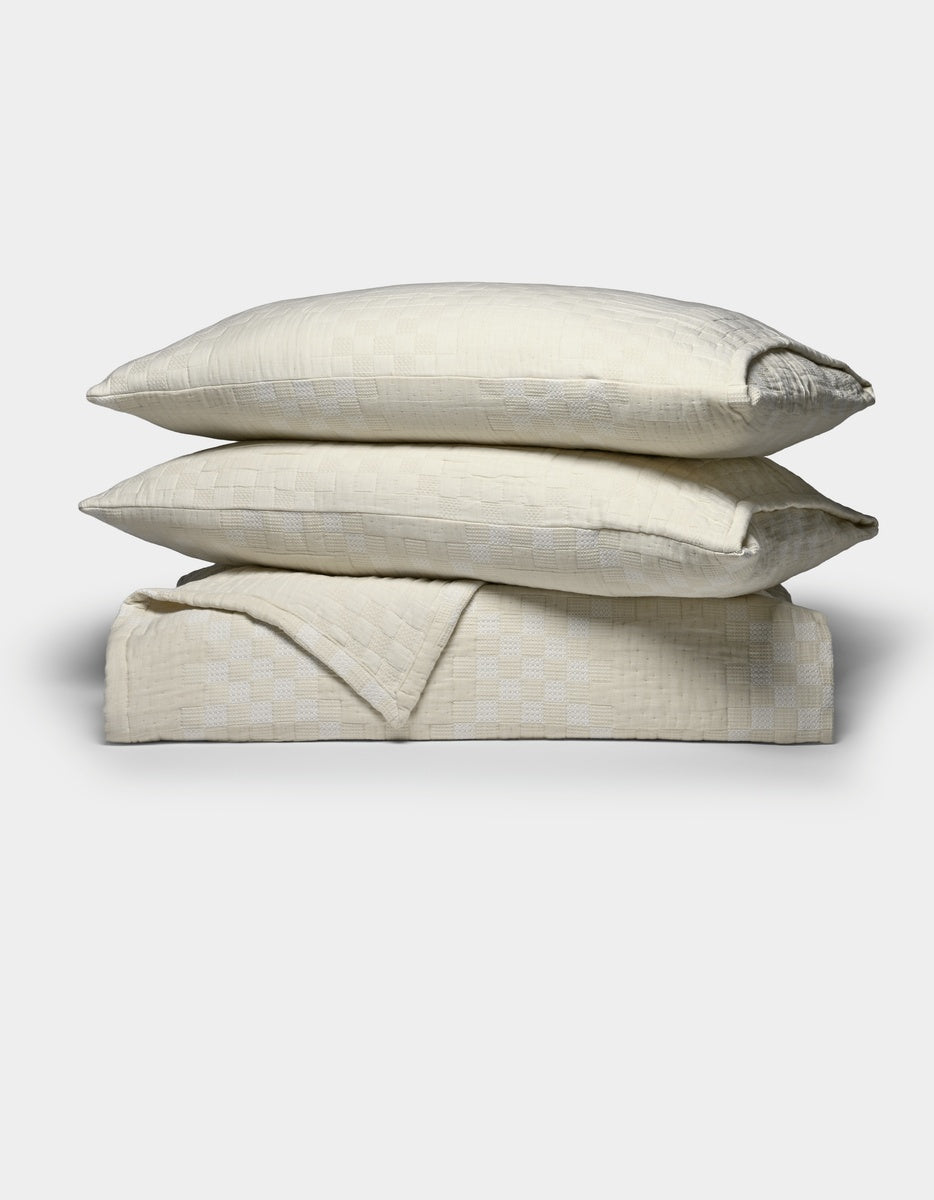 Two cream-colored pillows from the Cozy Earth Sonoma Stitch Quilt Bundle are stacked atop a matching folded blanket, all featuring a subtle checkered pattern, set against a plain white background.|Color:Vintage White