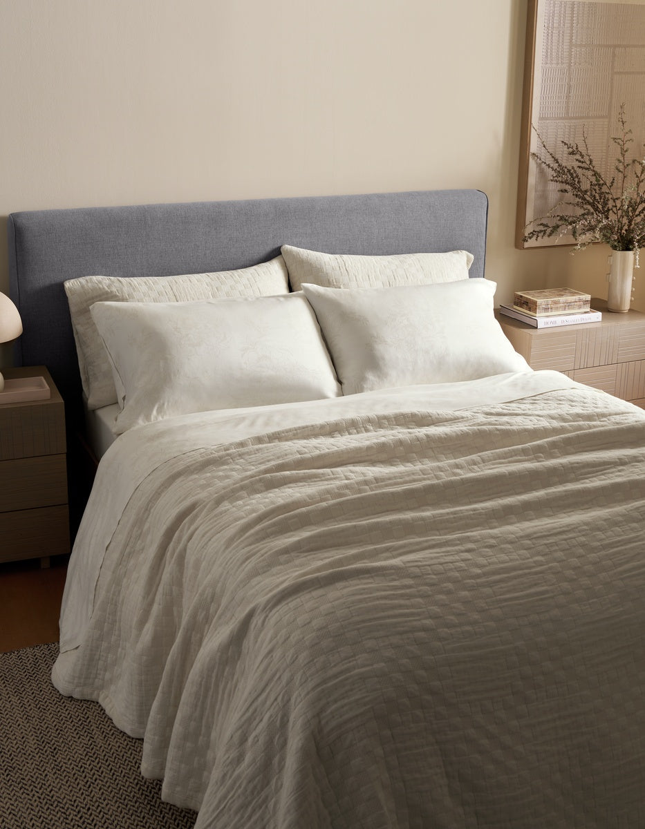A neatly made bed features the Cozy Earth Sonoma Stitch Quilt in cream, paired with white pillows and a gray upholstered headboard. Bedside tables display a lamp, books, and a vase with branches, while the room is styled in neutral tones with soft lighting.