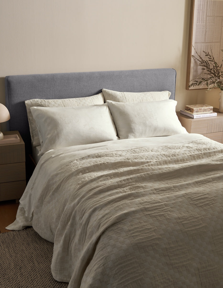 A neatly made bed features the Cozy Earth Sonoma Stitch Quilt in creamy tones with a gray upholstered headboard, flanked by bedside tables holding a lamp, books, and a vase of dried flowers in a softly lit, minimalist bedroom.