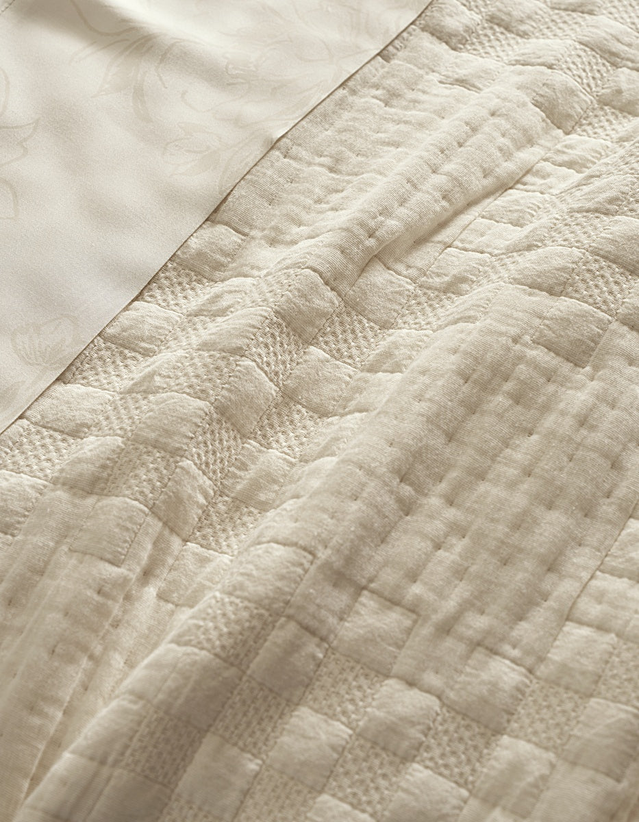 Close-up of the Cozy Earth Sonoma Stitch Quilt, featuring a cream-colored patchwork design with textured patterns, shown beside a smooth fabric adorned with a subtle floral print.