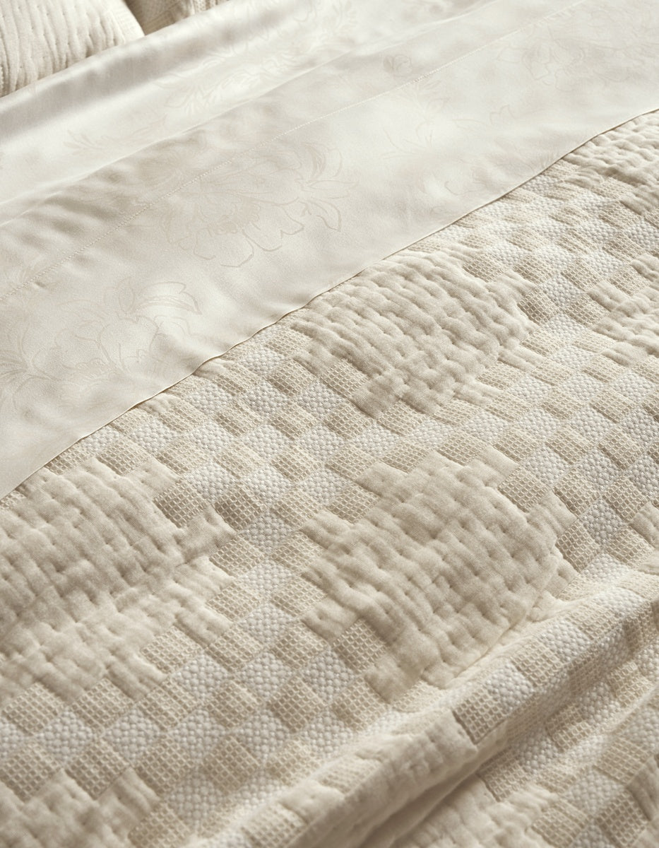A close-up of the Sonoma Stitch Quilt by Cozy Earth, showcasing its cream-colored fabric with a woven geometric and floral pattern, layered beneath a silky, floral-embossed sheet—both in soft, neutral tones.