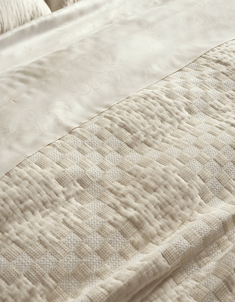 A close-up of the Sonoma Stitch Quilt by Cozy Earth, showcasing its cream-colored fabric with a woven geometric and floral pattern, layered beneath a silky, floral-embossed sheet—both in soft, neutral tones.