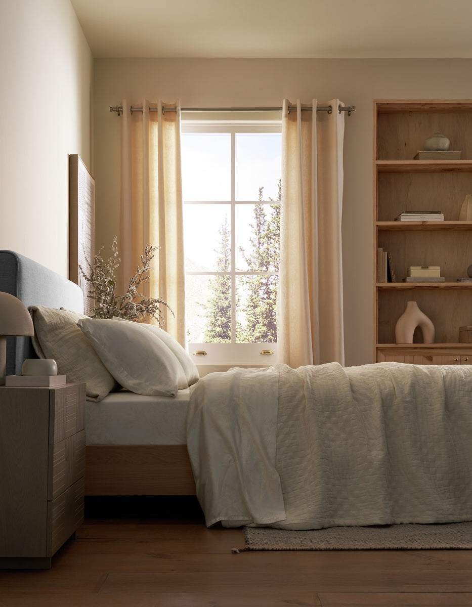 A sunlit bedroom with a made bed dressed in Cozy Earth’s Sonoma Stitch Quilt, beige bedding, a bedside table with a lamp, wooden shelves, and light curtains framing a window with trees visible outside.