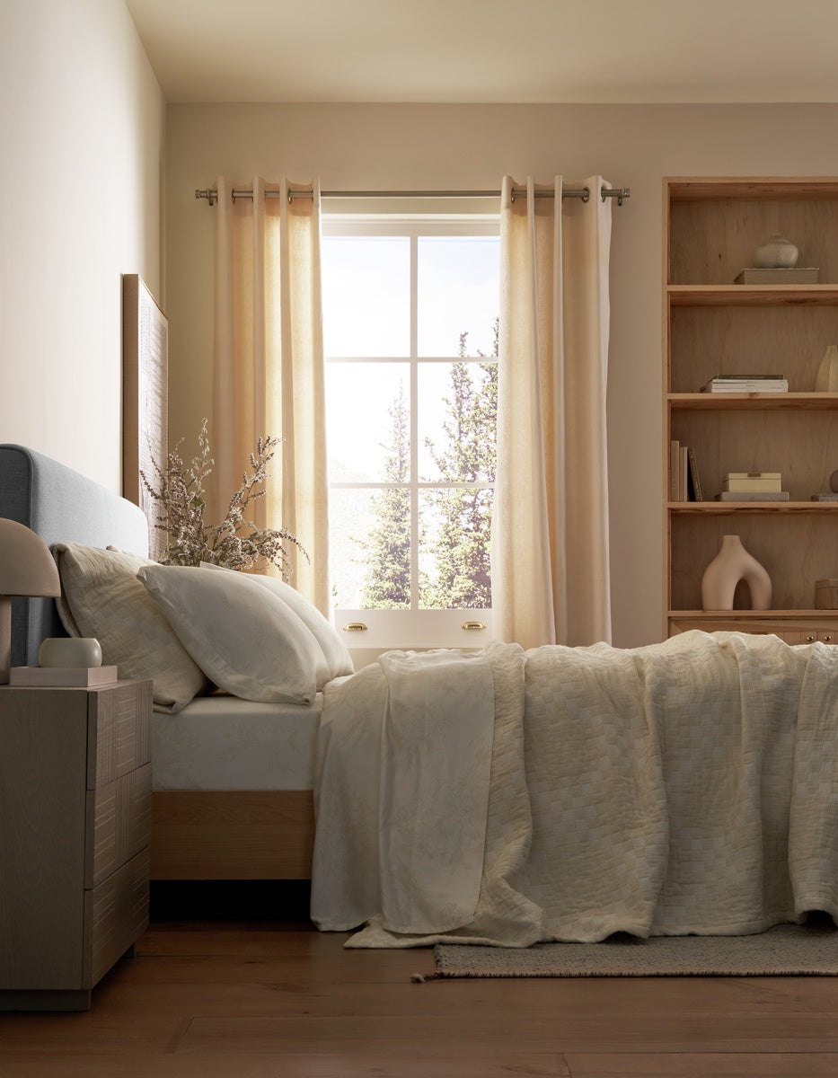 A neatly made bed with the Cozy Earth Sonoma Stitch Quilt in cream is positioned beside a window dressed in sheer curtains. Sunlight fills the room, highlighting a wooden nightstand and built-in shelving adorned with decorative objects.
